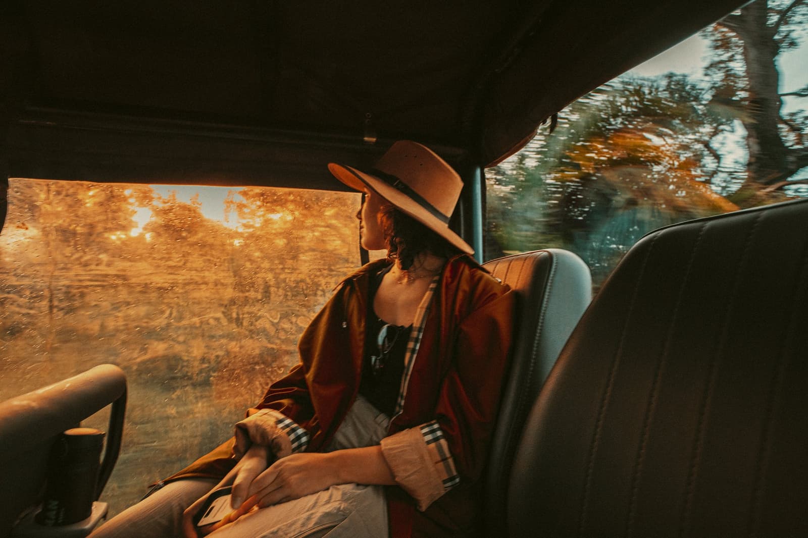 The light does something different out here, and somehow you always catch it from the back seat. A traveller in a wide-brim hat and rust-coloured wrap gazes out a rain-speckled jeep window at golden-hour light filtering through trees during a pantanal luxury safari transfer