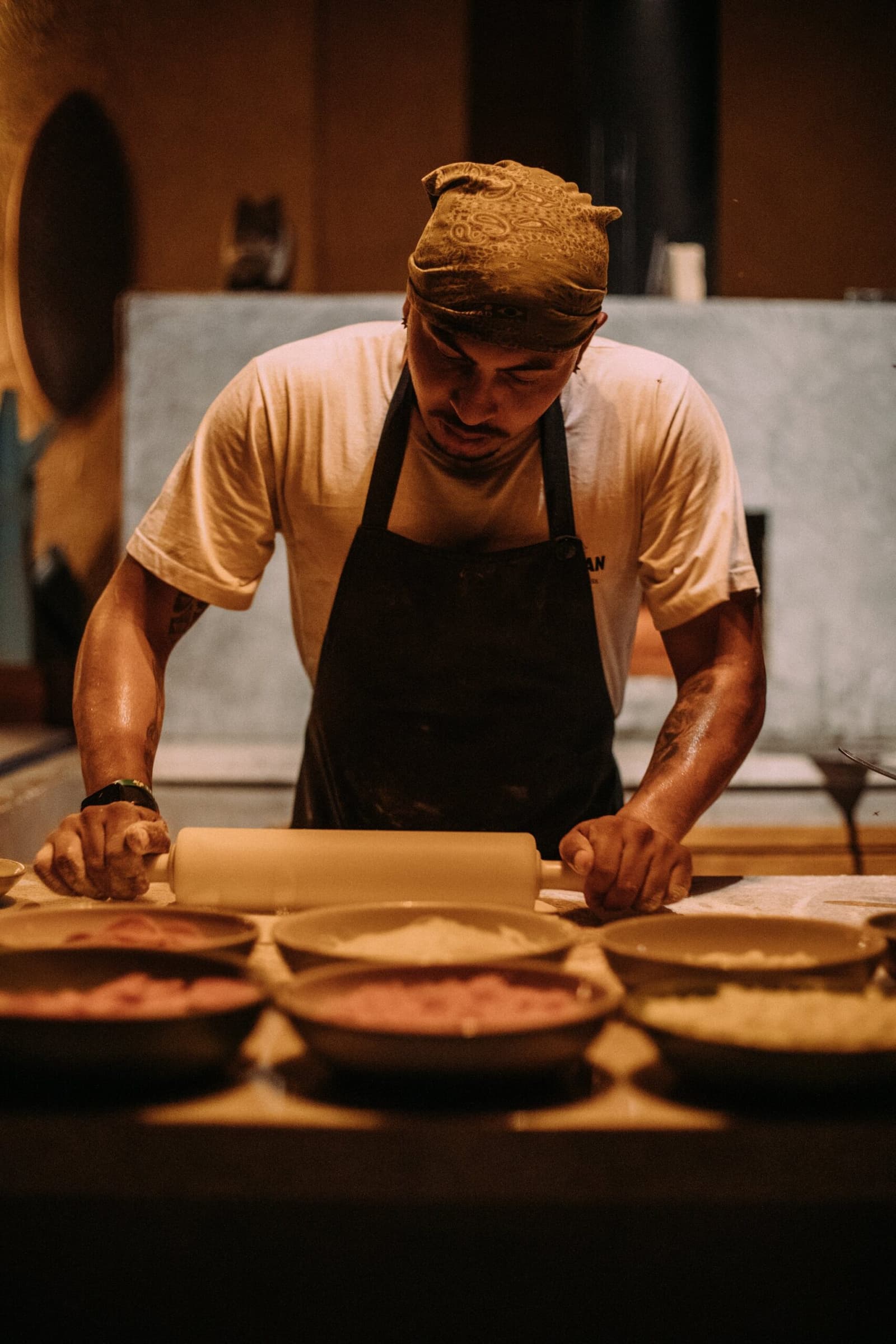 Good food in the middle of nowhere takes this much focus. A chef wearing a dark apron and bandana rolls out dough at a wooden prep station lined with ceramic bowls of filling, part of the pantanal luxury safari dining experience at Caiman