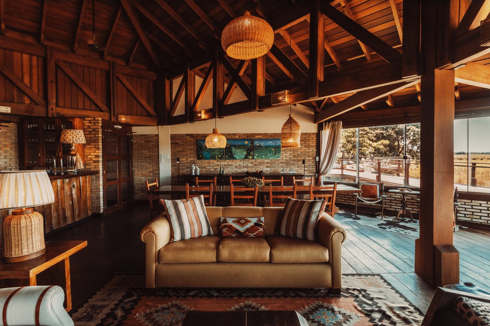 Cold drink in hand, savanna stretching out the window. This is the easy part of the day. Warmly lit common area of a pantanal luxury safari lodge, featuring a timber-framed vaulted ceiling with wicker pendant lights, a camel sofa with woven cushions, kilim rugs, and floor-to-ceiling windows opening onto savanna wetlands beyond