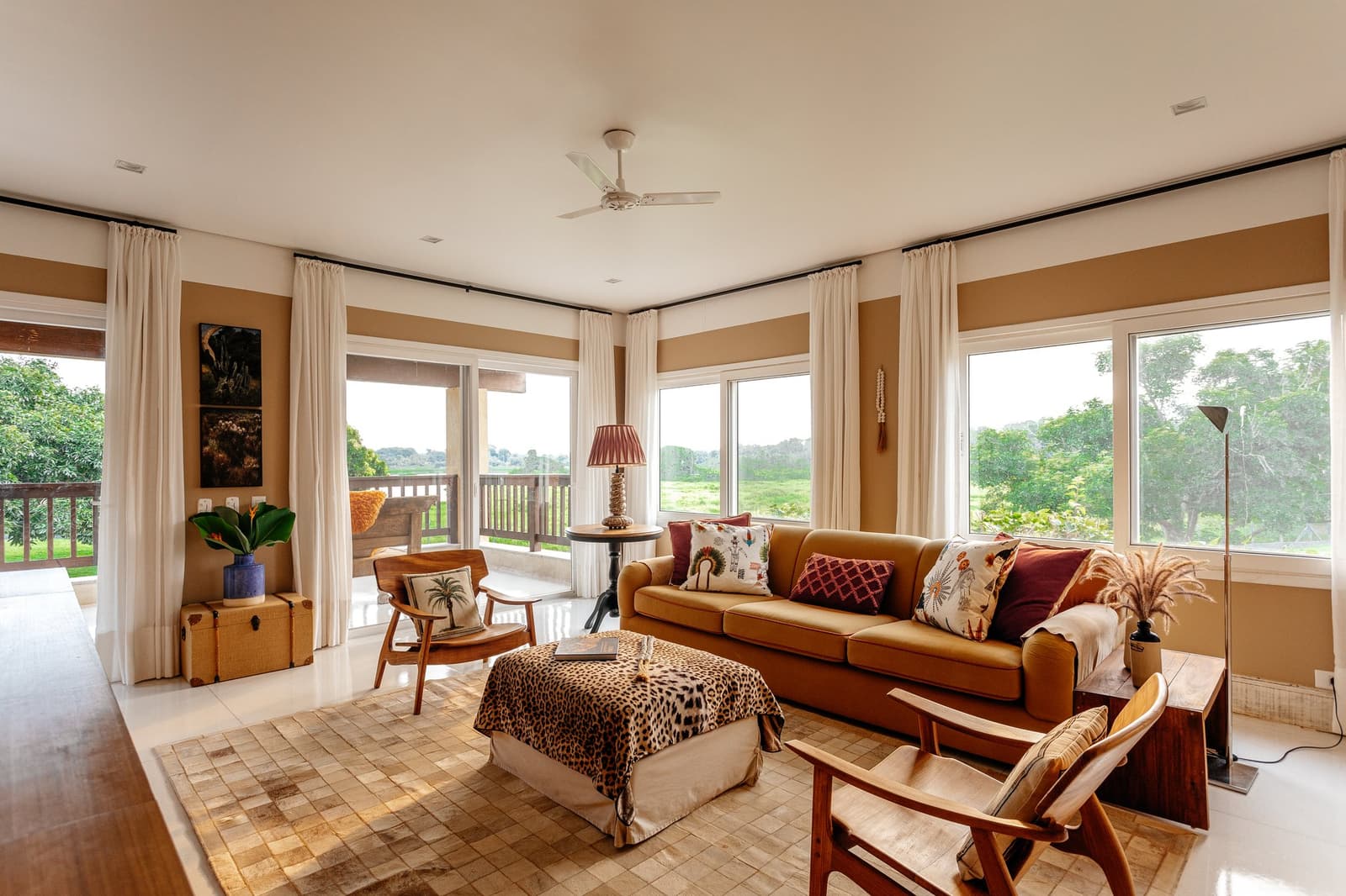 Comfortable enough that leaving for the morning game drive takes real willpower. Sunlit living room at a pantanal luxury safari lodge, featuring a caramel sofa with patterned cushions, a leopard-print ottoman, timber armchairs, and panoramic windows overlooking open wetland vegetation