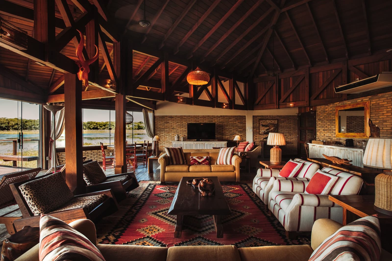 Come back from a wildlife walk and sink into this. The wetlands can wait five minutes. Spacious pantanal luxury safari lodge lounge with soaring timber-beam ceiling, kilim rug, animal-print cushions, and floor-to-ceiling glass doors opening onto a waterway view