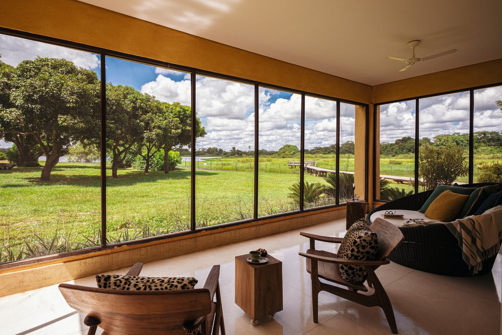 Your seat for watching the wetlands do their thing, no binoculars required. Elegant lounge area with floor-to-ceiling windows overlooking a green wetland meadow and wooden boardwalk at a pantanal luxury safari lodge
