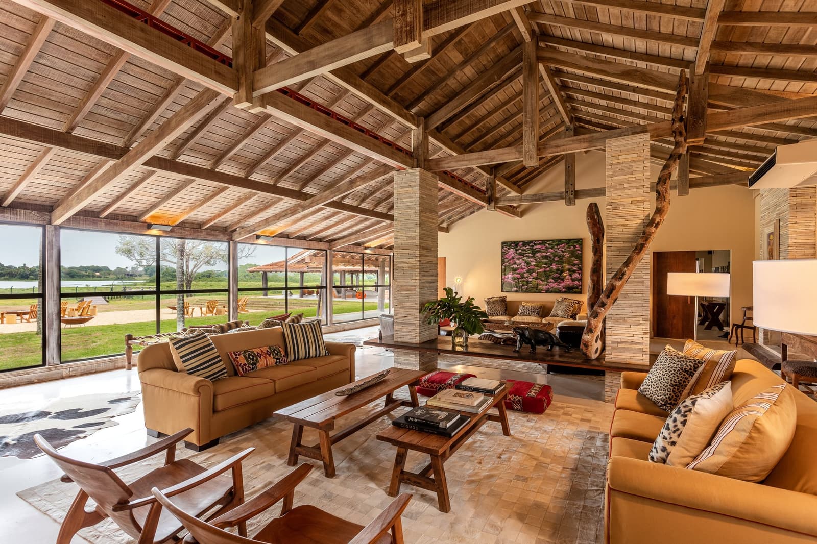 Rough timber, warm leather, and a view straight into the wild, camp life was never like this. Soaring timber-beam ceiling and ochre sofas in the open-plan lodge living room of a pantanal luxury safari property, with floor-to-ceiling glass walls framing green wetland views beyond