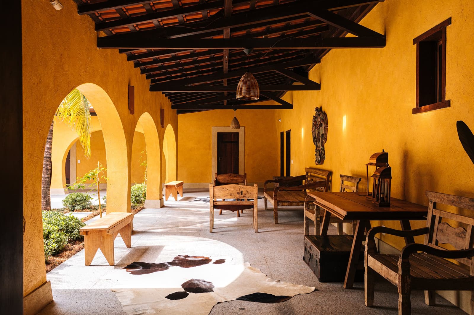 The kind of shaded corridor that makes you forget you had a schedule. Sunlit covered veranda of a pantanal luxury safari lodge, with golden ochre arched walls, dark timber beam ceiling, rustic wooden benches and dining table, wicker pendant lights, and a cowhide rug on the stone floor