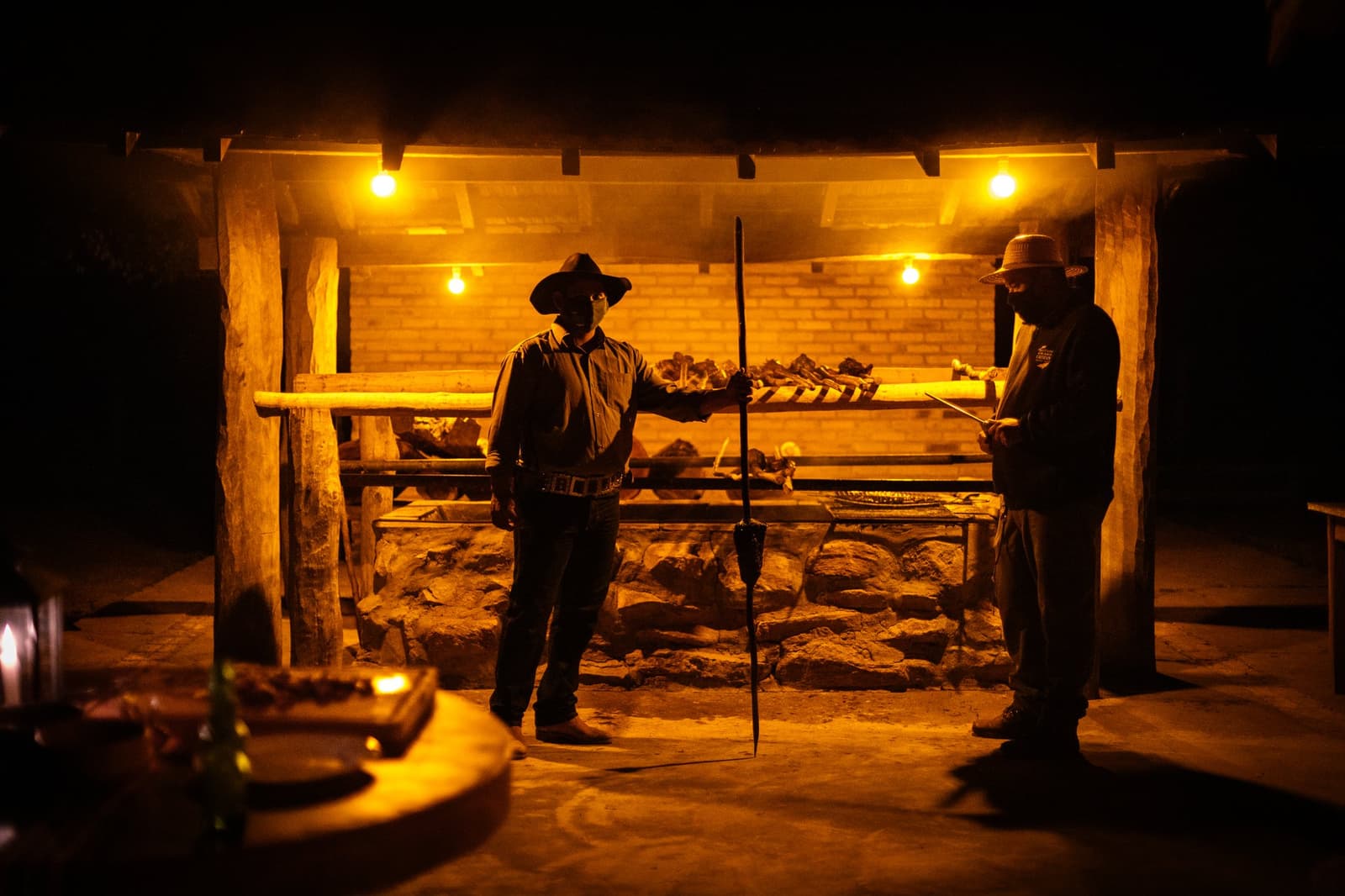 Dinner is serious business here, and these two have it handled. Two gauchos in wide-brimmed hats tend a large open-fire wood-spit grill glowing amber at night during a pantanal luxury safari dinner at Caiman