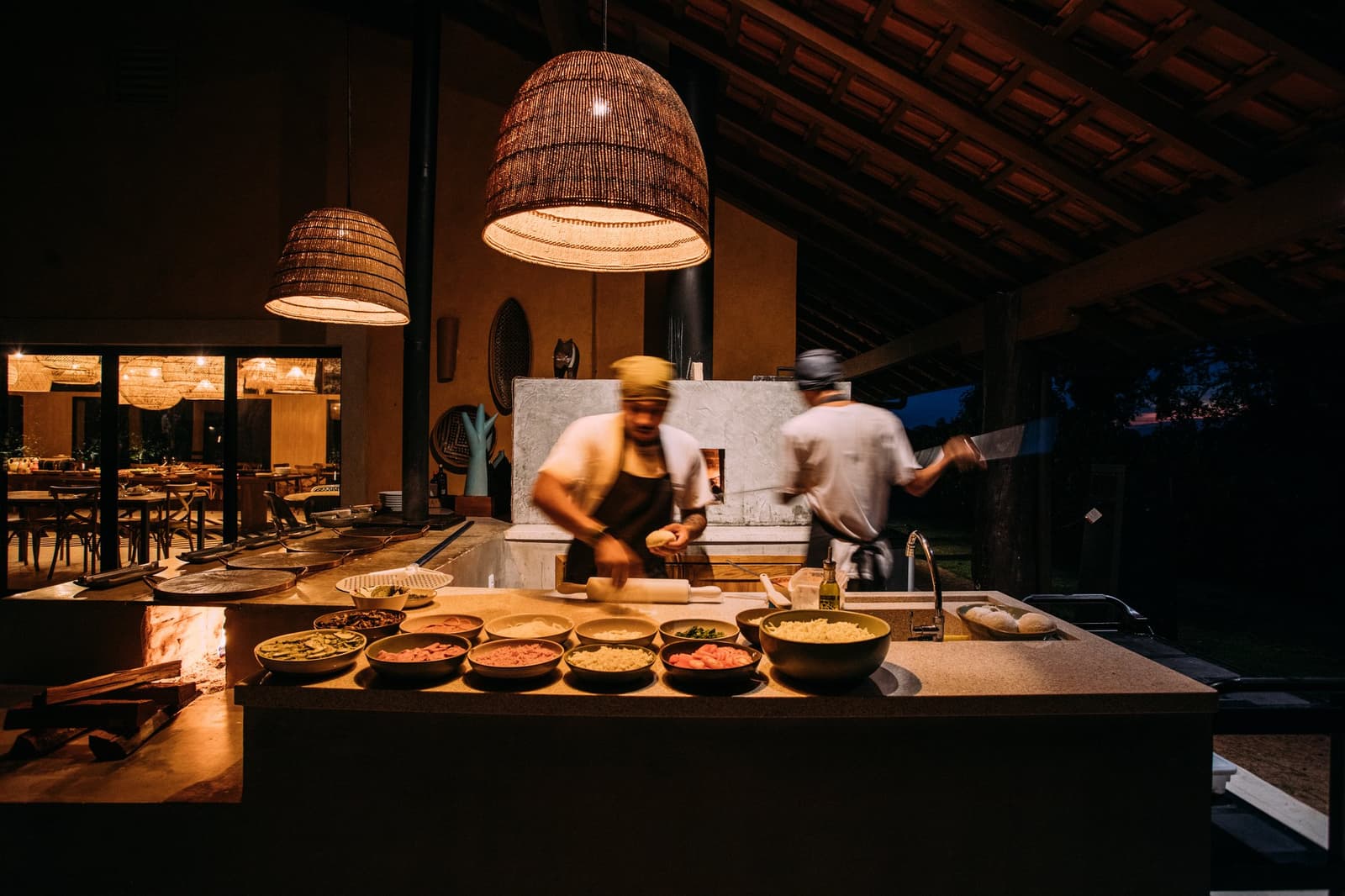 Dinner here is less a meal and more a small event worth staying up for. Two chefs in motion at an open-air wood-fired kitchen station during a pantanal luxury safari dinner, with bowls of fresh ingredients lit by warm wicker pendant lamps