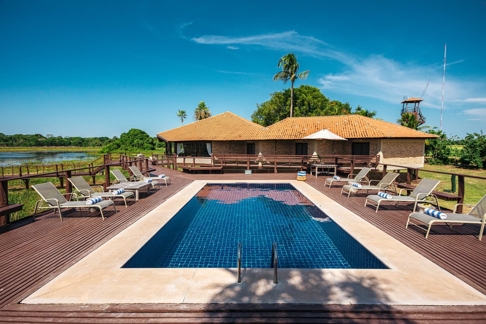 Cold water, warm afternoon, wetlands just past the fence. A tiled swimming pool on a hardwood deck surrounded by sun loungers, with a terracotta-roofed lodge and wetland panorama behind, at a pantanal luxury safari retreat
