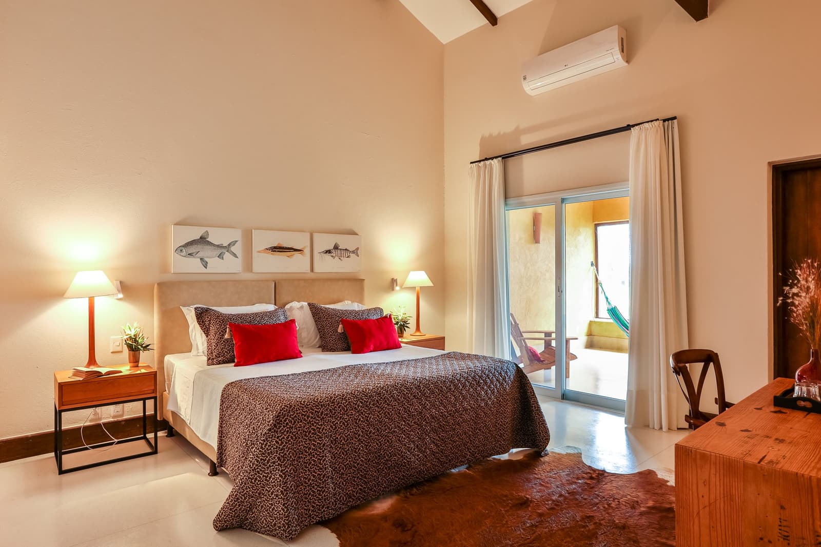 Fish art on the wall, a hammock just steps away, someone thought this through. Spacious pantanal luxury safari suite with a king bed dressed in leopard-print throws and red cushions, fish artwork above the headboard, vaulted timber ceiling, and a private terrace with a hammock visible through sliding glass doors