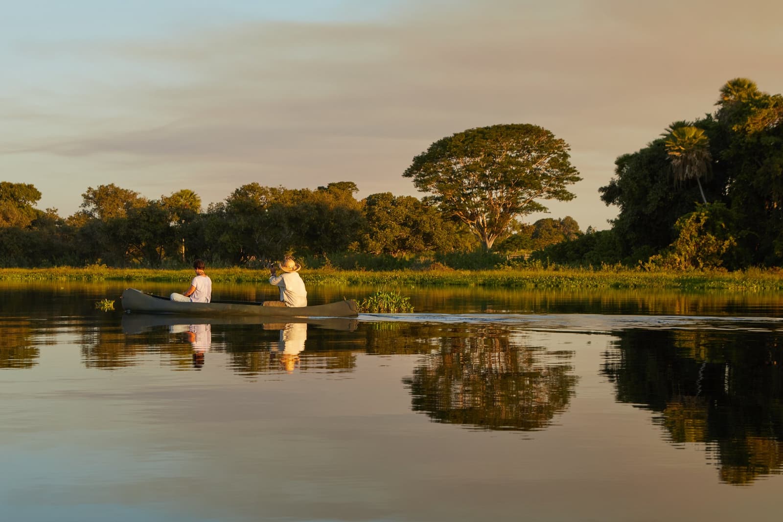 The quietest way to meet the Pantanal is at water level, at dusk. Two guests drifting in a canoe on glassy water at golden hour during a pantanal luxury safari, surrounded by lush wetland vegetation and large trees reflected in the still river