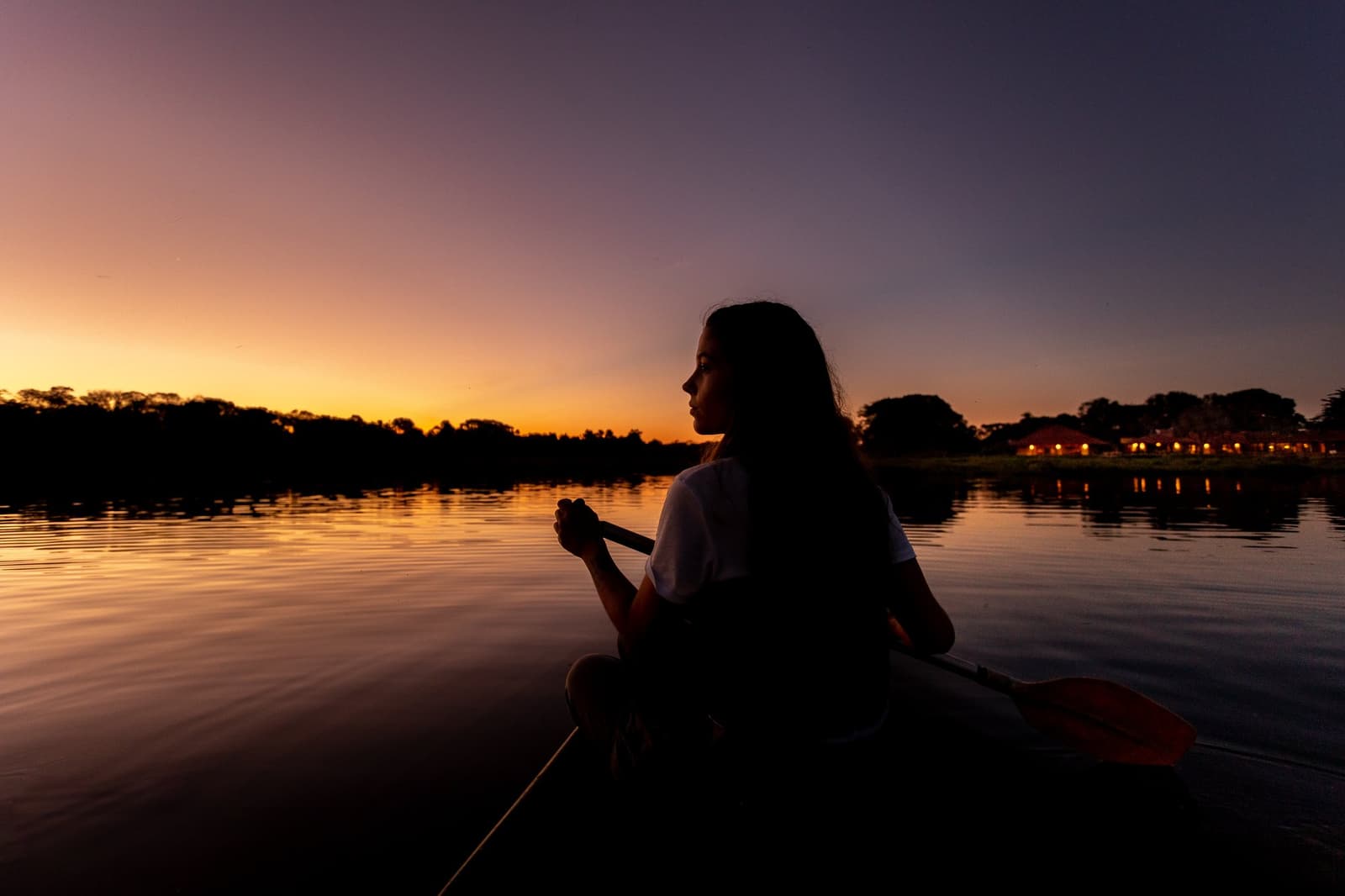 The Pantanal slows time down, and on the water at dusk, you really feel it. Silhouette of a woman holding a canoe paddle on a glassy river during a pantanal luxury safari, with a vivid orange and purple sunset reflected on the water and lodge lights glowing on the far bank
