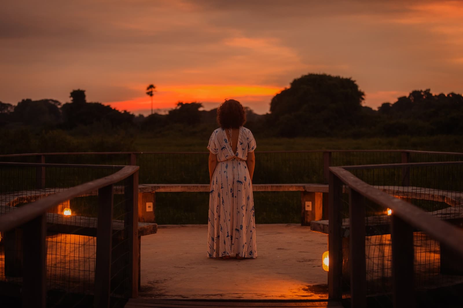 Some sunsets demand you put the camera down and just watch. A woman in a floral maxi dress stands on a candlelit wooden deck, watching a blazing orange sunset over open wetland flats on a pantanal luxury safari