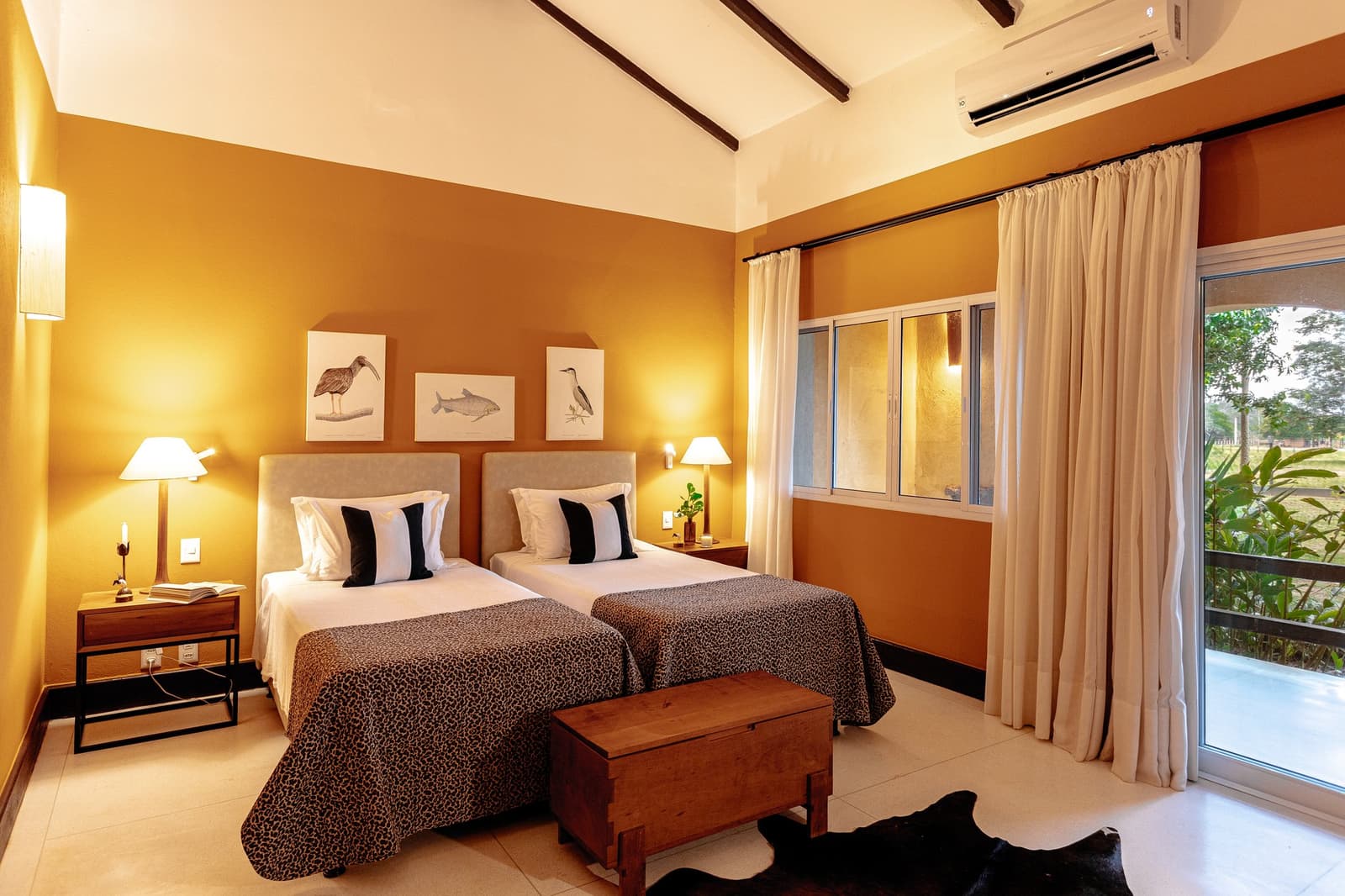 Nature prints on the wall, the wetlands just outside the door. Twin bedroom at a pantanal luxury safari lodge featuring warm ochre walls, wildlife botanical prints, leopard-print bedspreads, exposed timber ceiling beams, and a sliding glass door opening onto lush greenery