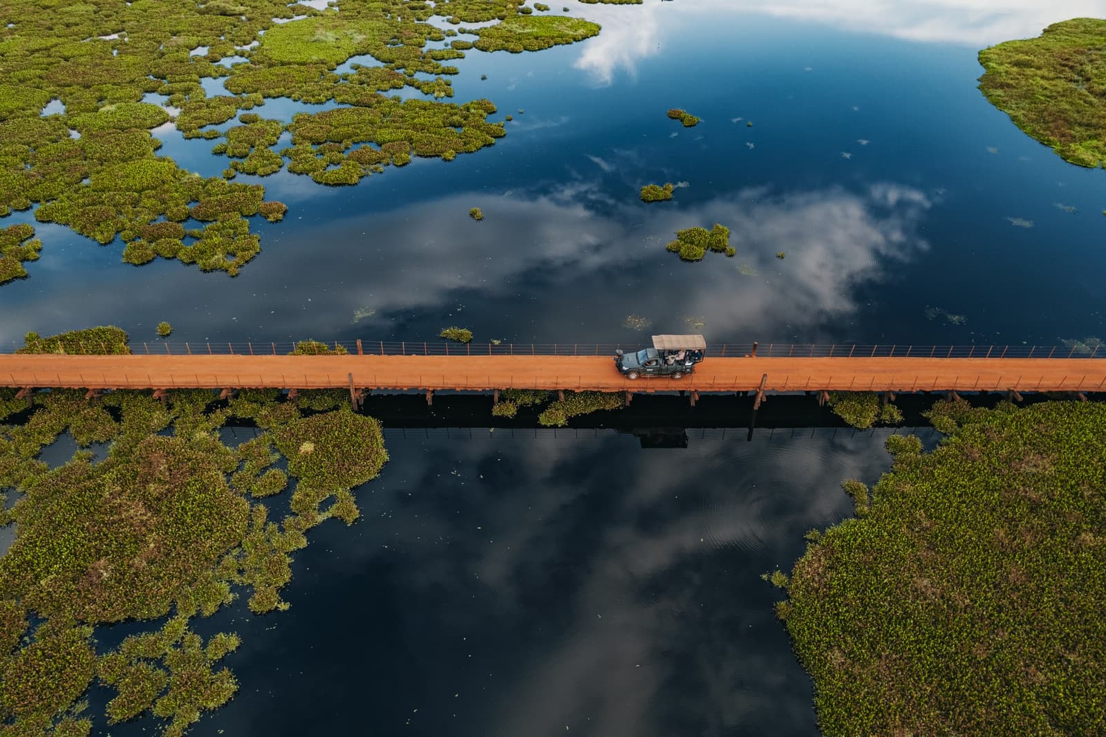 The bridge cuts the wetland in two, and the clouds can't decide which side they prefer. Aerial view of a safari vehicle crossing a red-earth bridge over mirror-still flooded wetlands scattered with green aquatic vegetation during a pantanal luxury safari