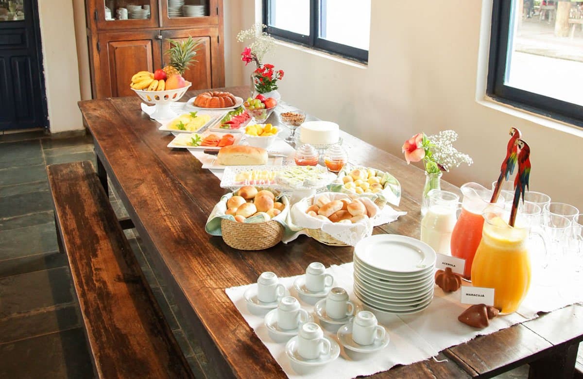 Fresh fruit, warm bread, and two ceramic macaws judging your portion size. A generous Brazilian breakfast buffet laid out on a long wooden table at a Pantanal tour from Campo Grande lodge, featuring fresh tropical fruits, bread rolls, cakes, acerola and maracujá juices, and small espresso cups
