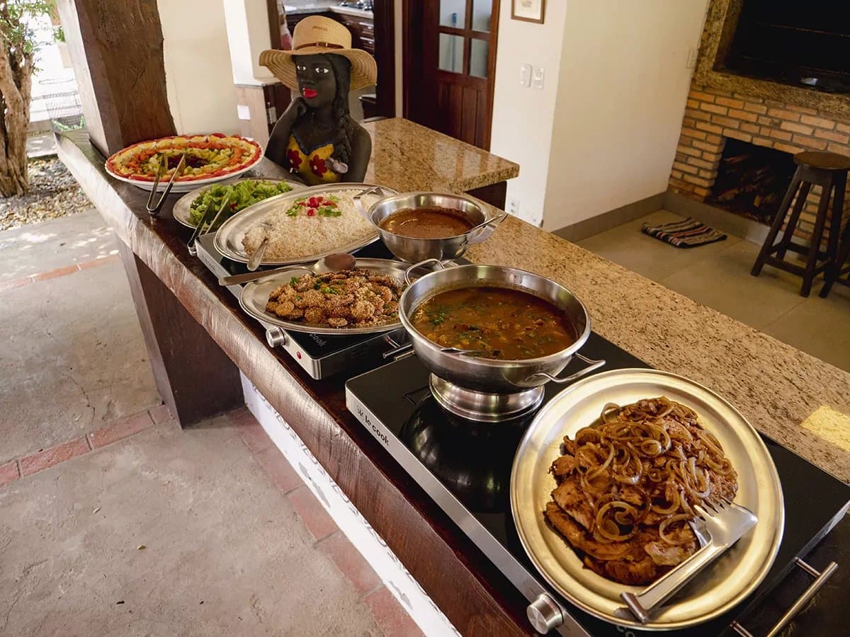 Fuel up well, the caimans aren't waiting for anyone. A generous lodge buffet spread on a granite counter featuring grilled meat with caramelised onions, two warm stews, rice, a fried side dish, green salad, and a colourful fruit platter — part of the full-board experience on a pantanal tour from campo grande