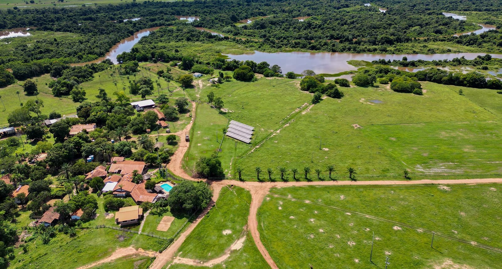 Life here is shaped by water, grass, and the slow curve of the river. Aerial view of a Pantanal ecolodge on a pantanal tour from Campo Grande, with solar panels, red-roofed buildings, a swimming pool, dirt tracks, green pastures, and a winding river visible in the background