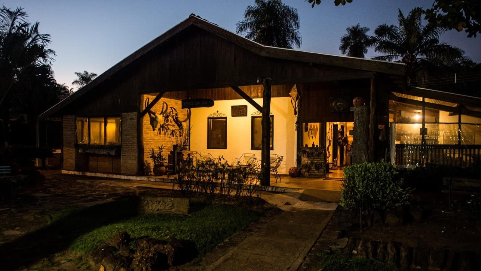 After a long day in the wild, this is exactly what you want to see. A warmly lit rustic ecolodge with a wooden gabled roof and open veranda glowing with amber light at dusk, surrounded by palm trees and tropical gardens, on a pantanal tour from campo grande