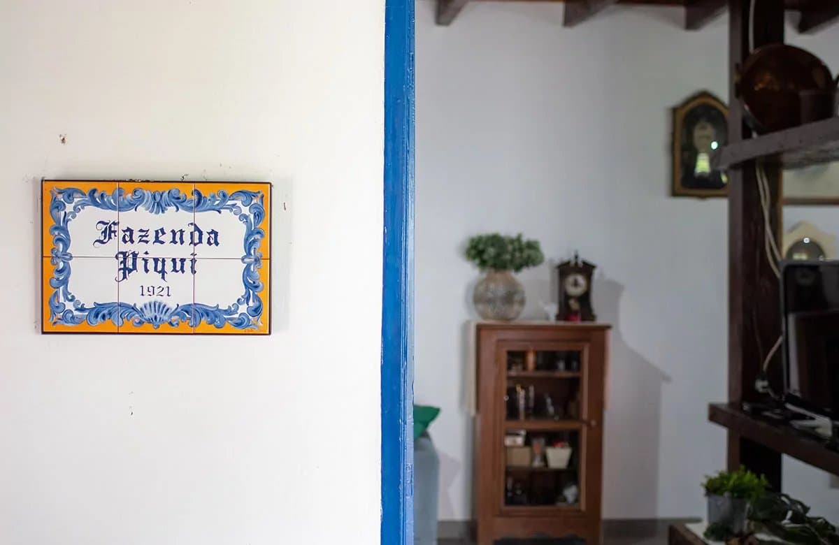 A farm with a founding date and a tile that means it. Hand-painted ceramic tile plaque reading 'Fazenda Piqui 1921' mounted on a white wall beside a blue-framed doorway leading into a rustic interior, at the start of a pantanal tour from campo grande