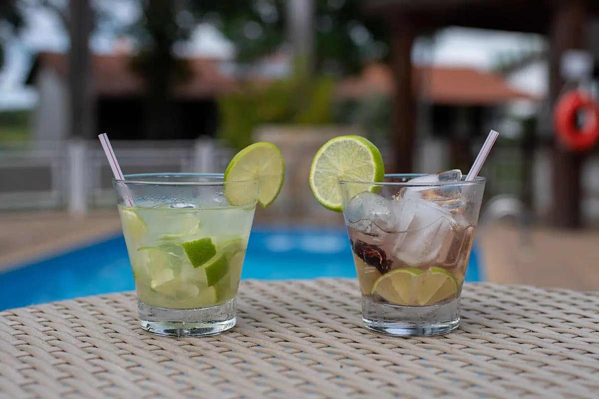 The afternoon belongs to whoever ordered first. Two lime-garnished cocktails with ice and straws on a wicker table beside a blue swimming pool at a Pantanal tour from Campo Grande ecolodge