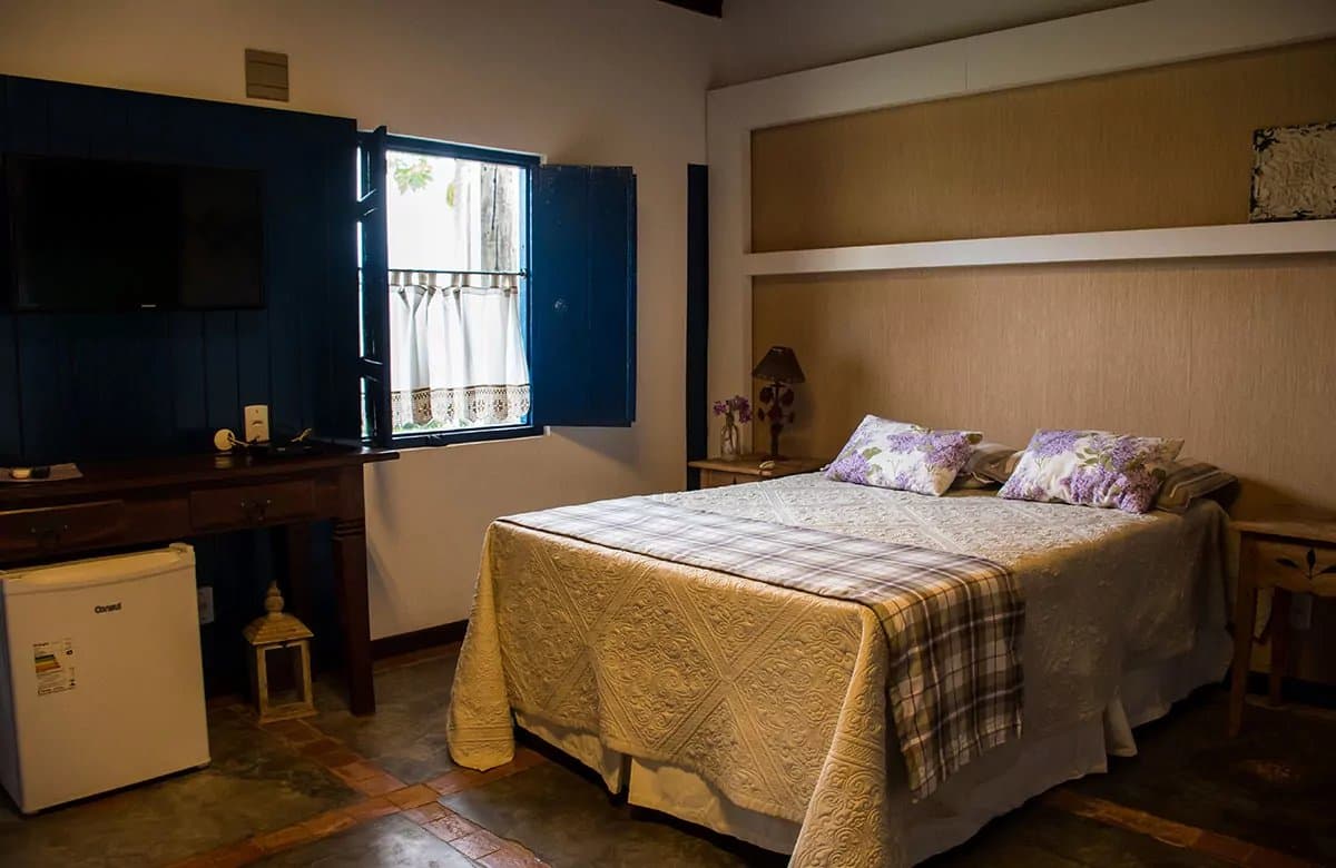 Simple, solid comfort after a long day in the field. A cozy double room on a pantanal tour from campo grande, featuring a quilted bedspread with floral pillows, blue wooden shutters open to garden greenery, a wooden desk with flat-screen TV, and a mini-fridge on terracotta tile floors