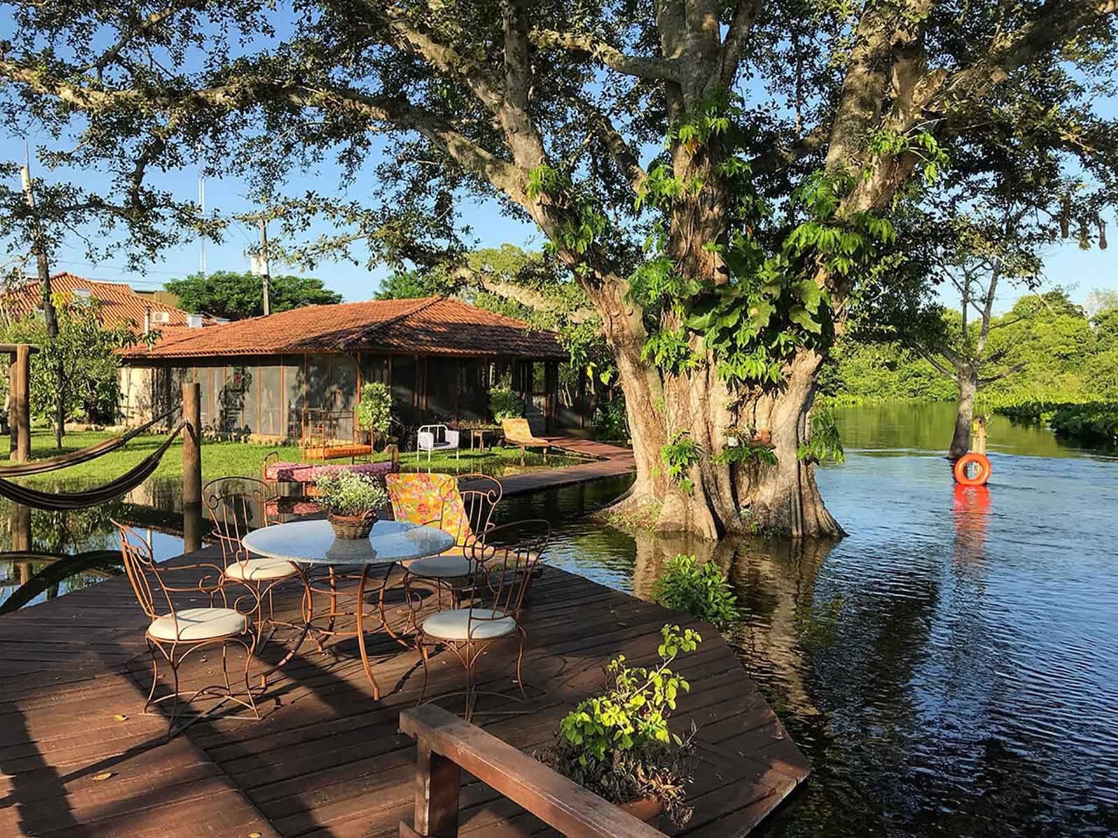 The kind of breakfast spot that makes leaving feel unreasonable. A wooden deck with wrought-iron café chairs overlooking calm river water, with a large tree growing from the bank and a terracotta-roofed ecolodge visible behind lush greenery — an idyllic setting on a pantanal tour from campo grande