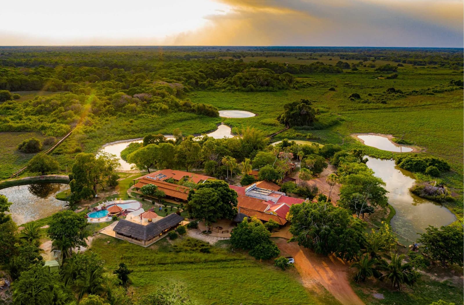 Your base camp happens to be one of the prettiest spots in the floodplain. Aerial view of a Pantanal eco lodge surrounded by winding lagoons, lush wetland vegetation, and a terracotta-roofed main building with a swimming pool, on a Pantanal Tour from Cuiabá at golden sunset