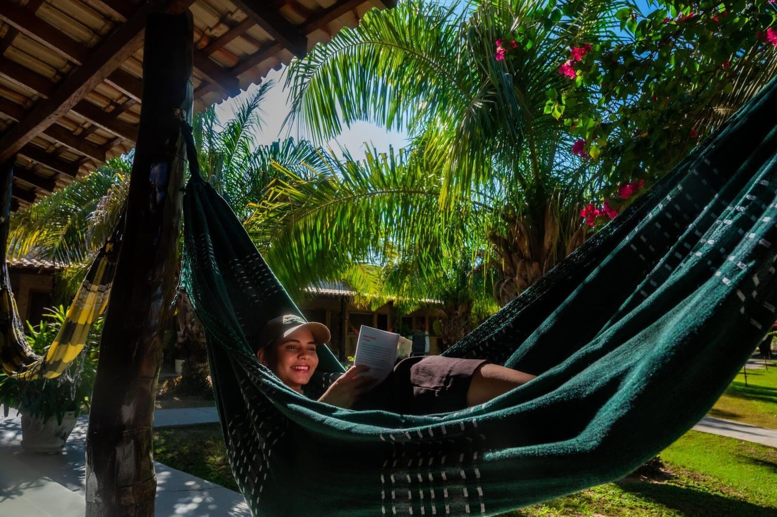 The book can wait. Actually, no it can't, and that's the point. A smiling traveller reads a book while resting in a dark green hammock on a shaded lodge veranda, surrounded by palm fronds and pink bougainvillea, during a Pantanal Tour from Cuiabá