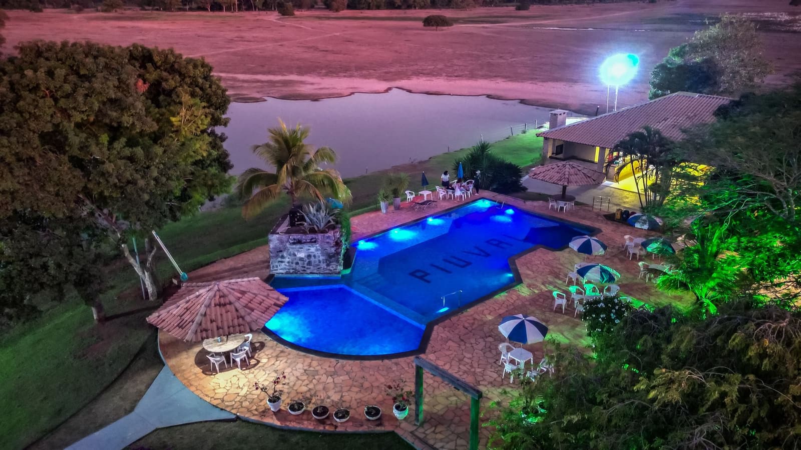 After a long day on the water, this pool earns every bit of its blue glow. Aerial night view of an illuminated swimming pool at a Pantanal Tour from Cuiabá lodge, with patio umbrellas, a stone water feature, and a natural lake stretching across open savanna in the background