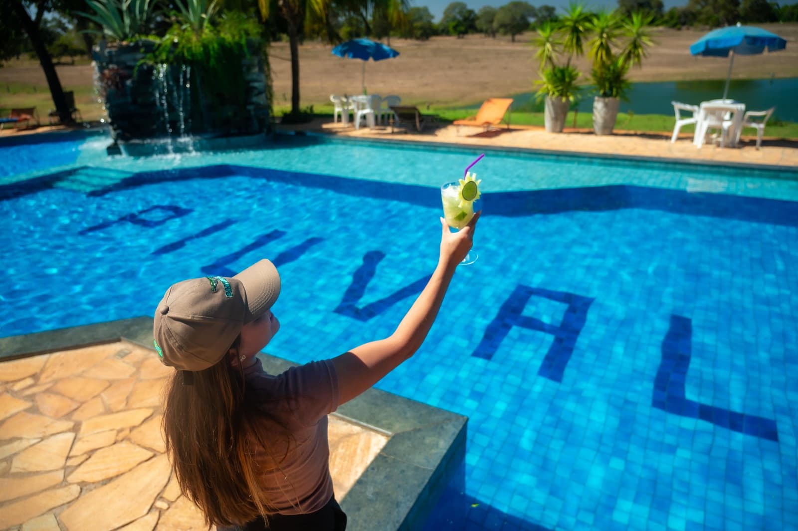 The lodge's way of saying: you made it, now relax. A guest raises a lime cocktail beside the blue tiled pool of a Pantanal tour from Cuiabá lodge, with sun loungers and a tranquil lake visible in the background