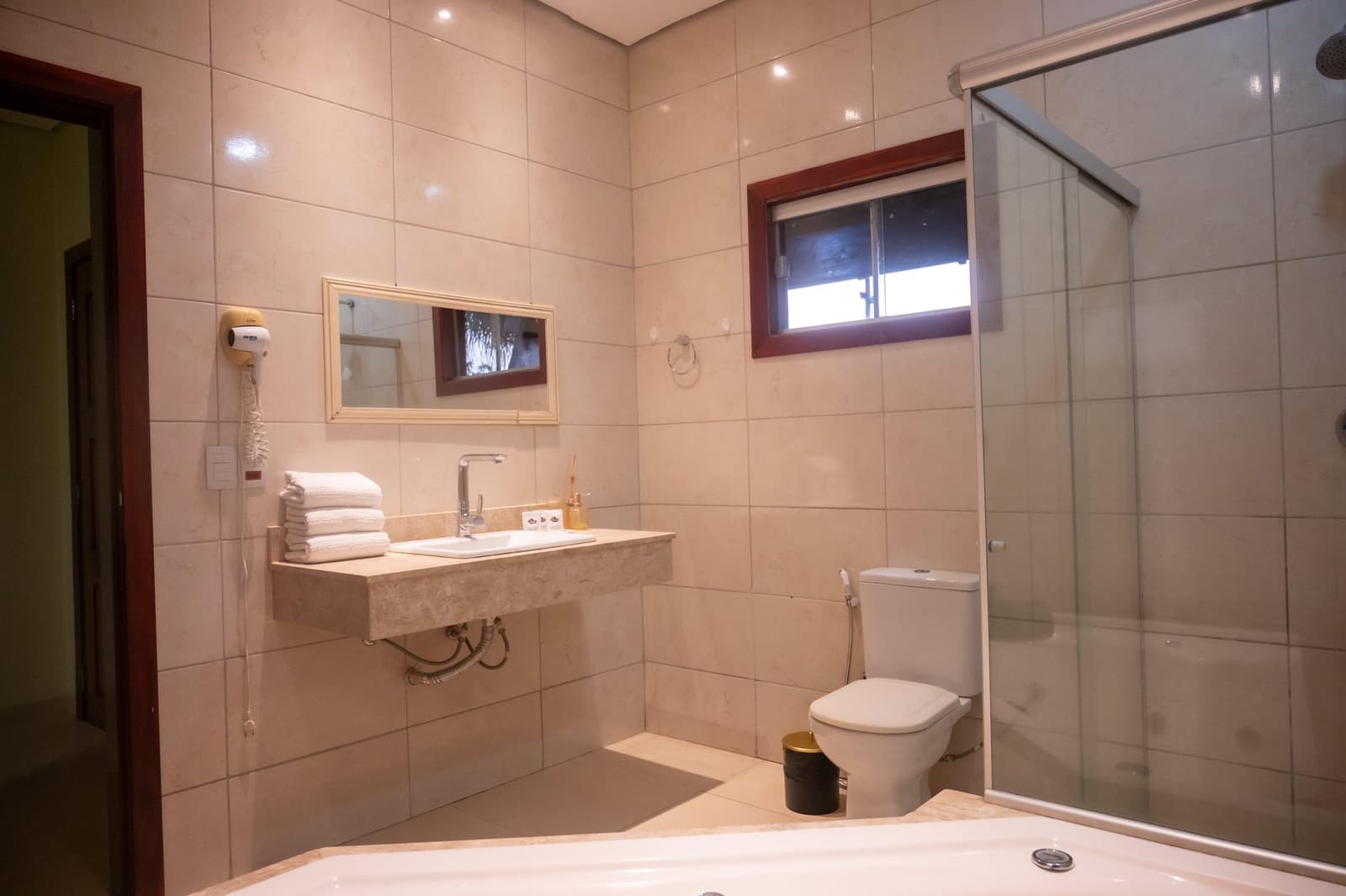 Warm tiles, fresh towels, and a hot shower waiting after a long day in the field. Spacious private bathroom at a Pantanal Tour from Cuiabá lodge, featuring a marble-finish vanity, folded white towels, glass-enclosed shower, bathtub, and a wall-mounted hair dryer