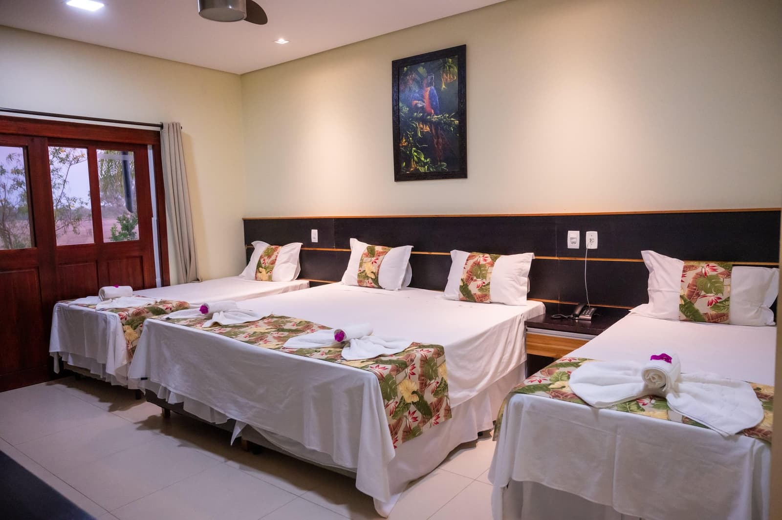 Clean, comfortable, and ready for you after a long day in the field. A triple-occupancy lodge room on a Pantanal Tour from Cuiabá, with three neatly made beds featuring tropical floral runners, rolled towels with purple orchid accents, and a framed macaw painting above the headboard