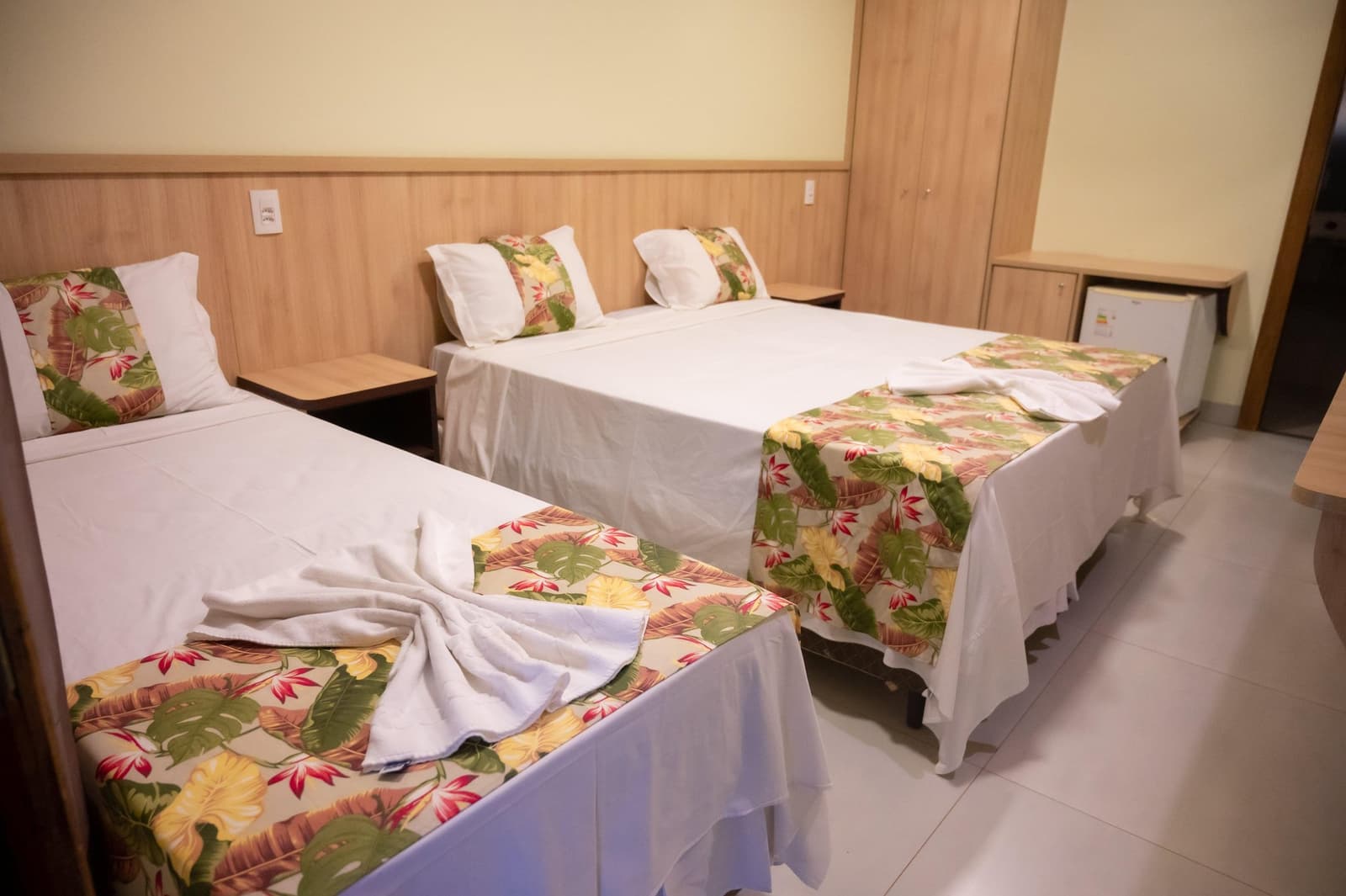 Neat beds, a folded towel, and a tropical print, someone here takes the welcome seriously. Three neatly made beds with tropical floral bedspreads and folded white towels in a clean, wood-panelled guest room on a Pantanal Tour from Cuiabá