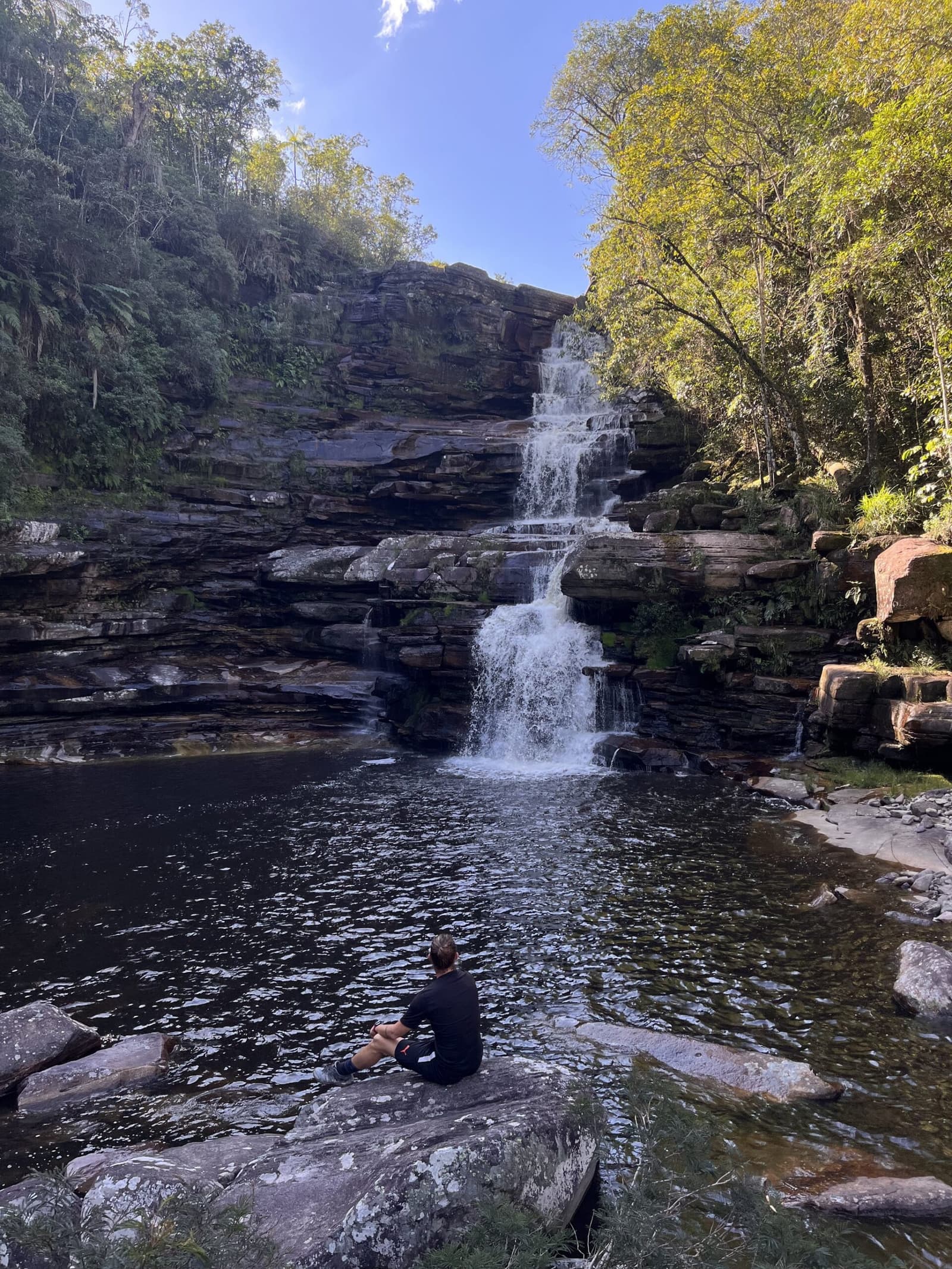Some moments are best taken sitting down, with no agenda. A hiker sits alone on a flat sandstone rock beside a dark natural pool, gazing up at a tiered waterfall cascading down layered rock faces during the Pati Valley hike, surrounded by lush green forest under a clear blue sky