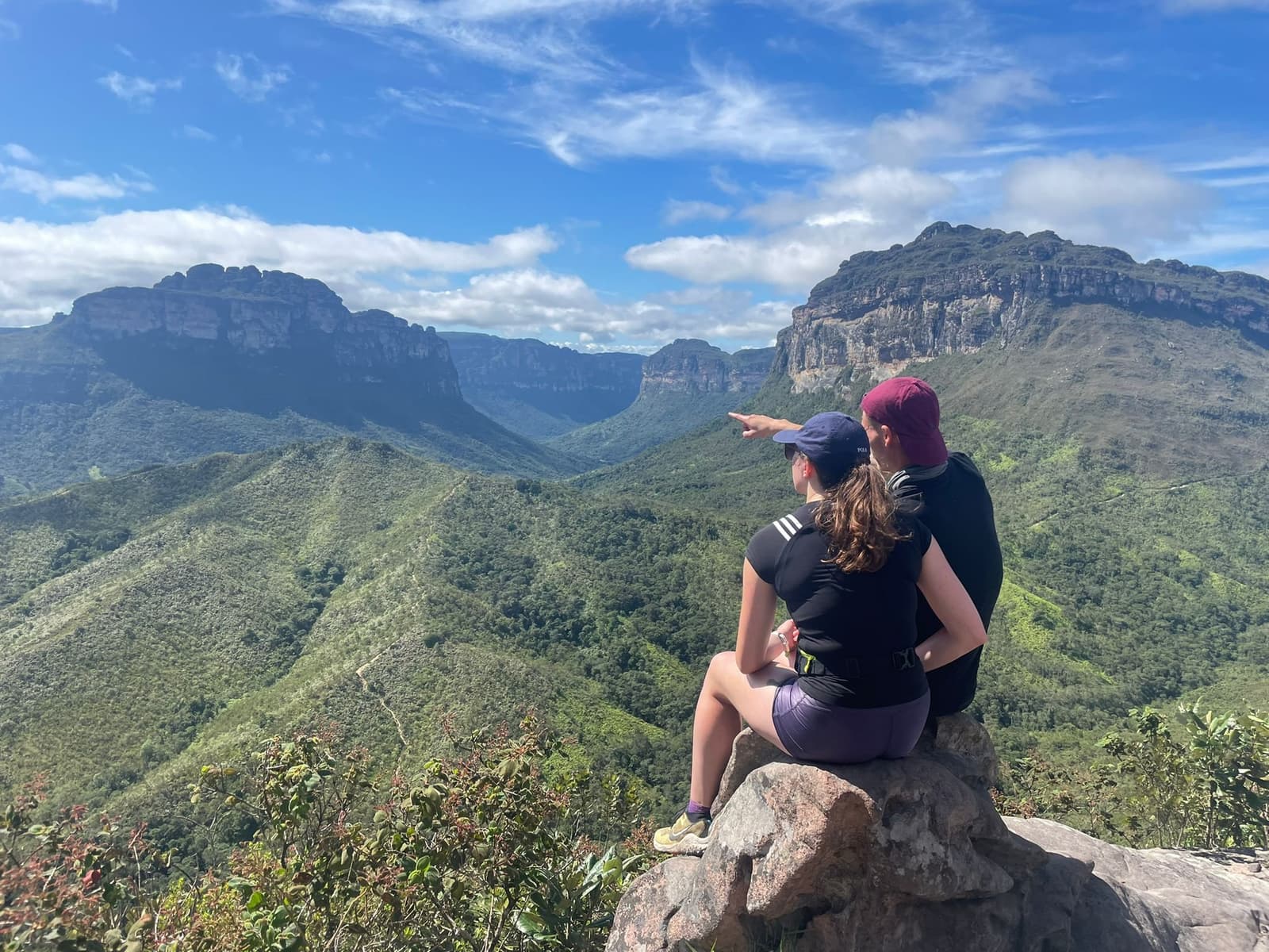 That moment when one of you spots something the other absolutely has to see. Two hikers sit on a rocky outcrop on the Pati Valley hike, one pointing toward the vast green canyon flanked by towering sandstone plateaus under a blue sky