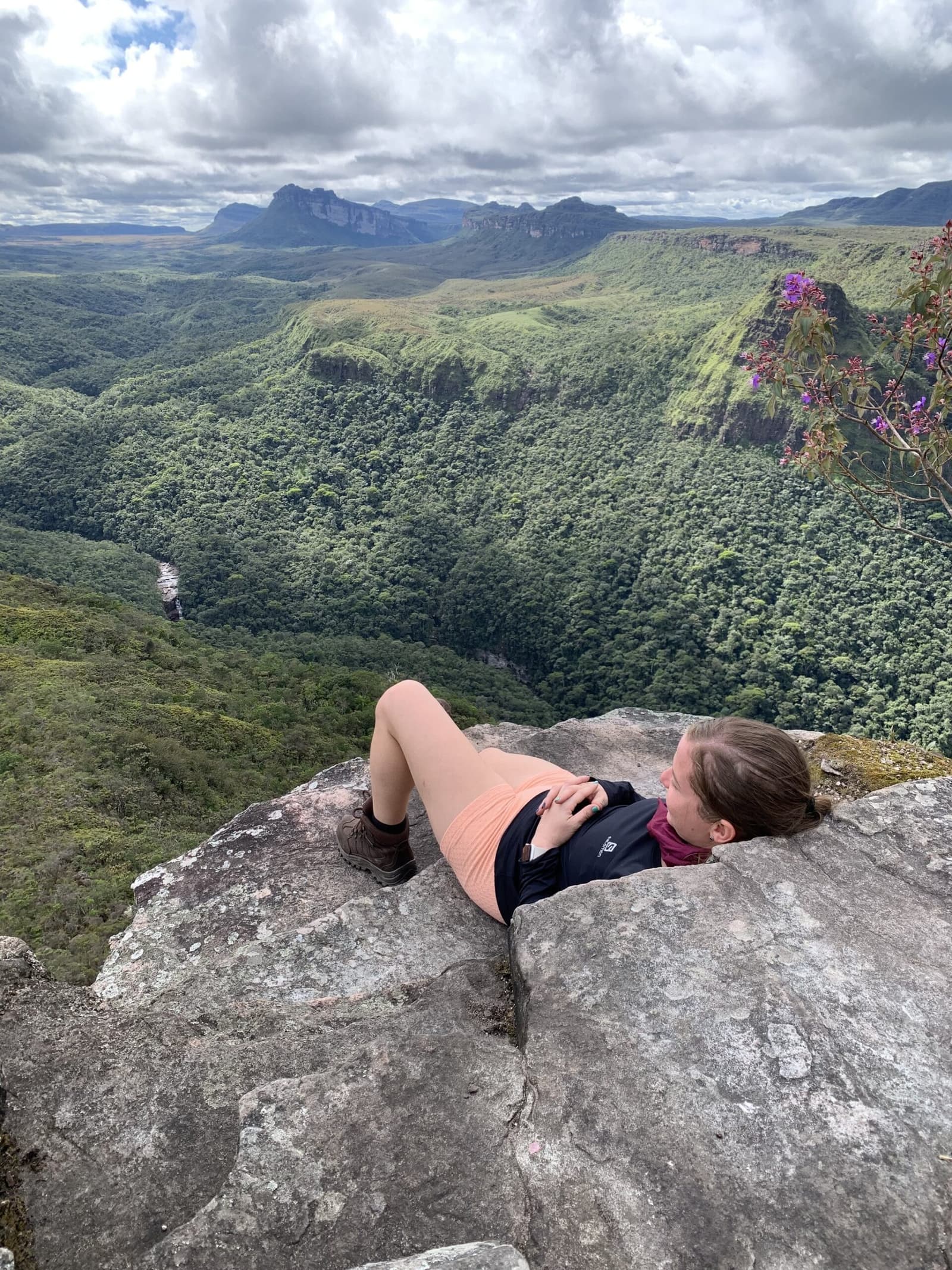 The best seat in the house, no reservation required. A hiker lies back on a rocky cliff edge on the Pati Valley hike, gazing over a deep green forested canyon with flat-topped sandstone mountains and a distant waterfall under a cloudy sky