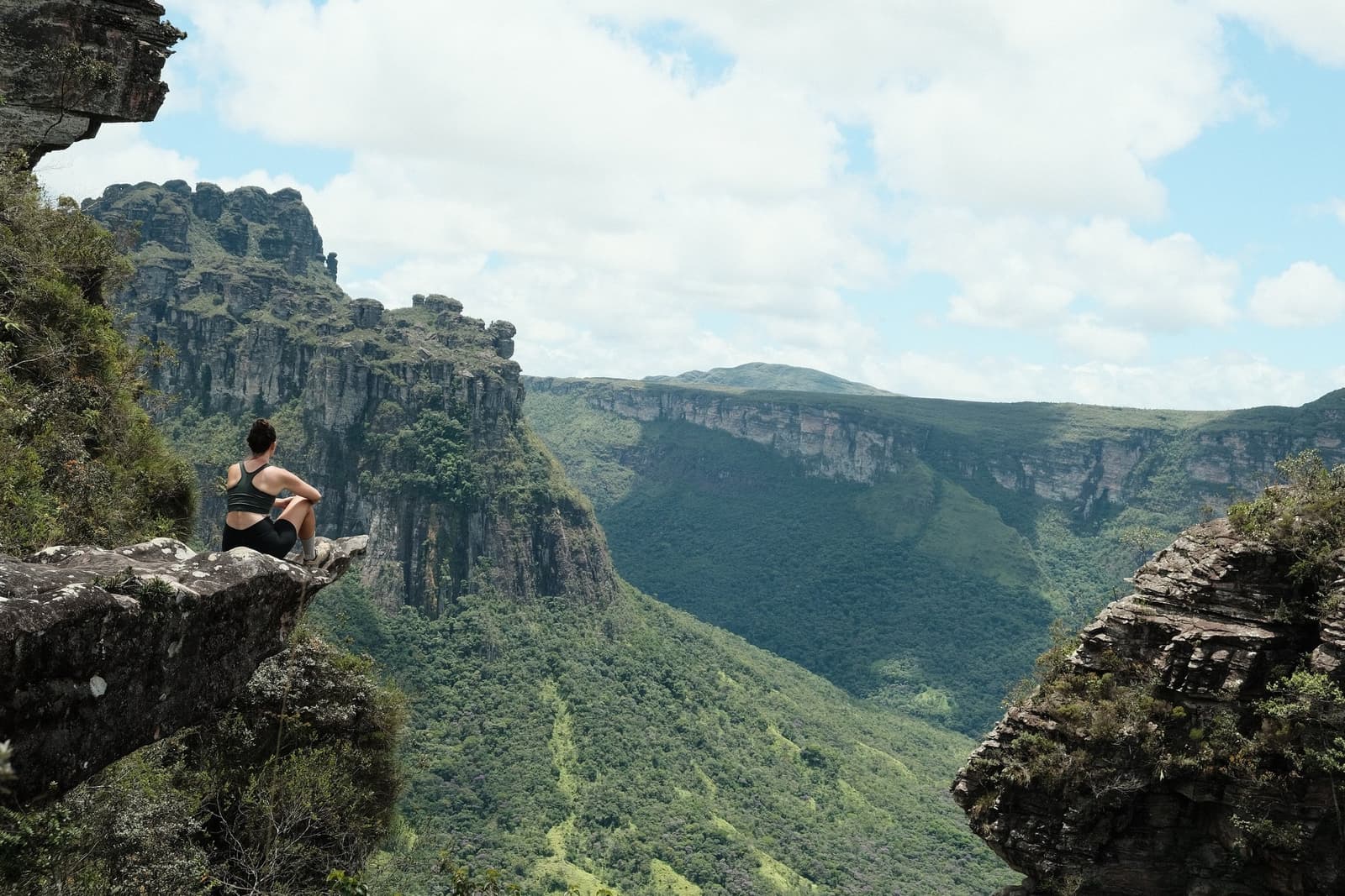 Some silences you have to earn with your legs. A hiker sits alone on a rock ledge overlooking a vast green canyon during the Pati Valley hike, with towering sandstone cliffs and forested valley walls stretching into the distance under a partly cloudy sky