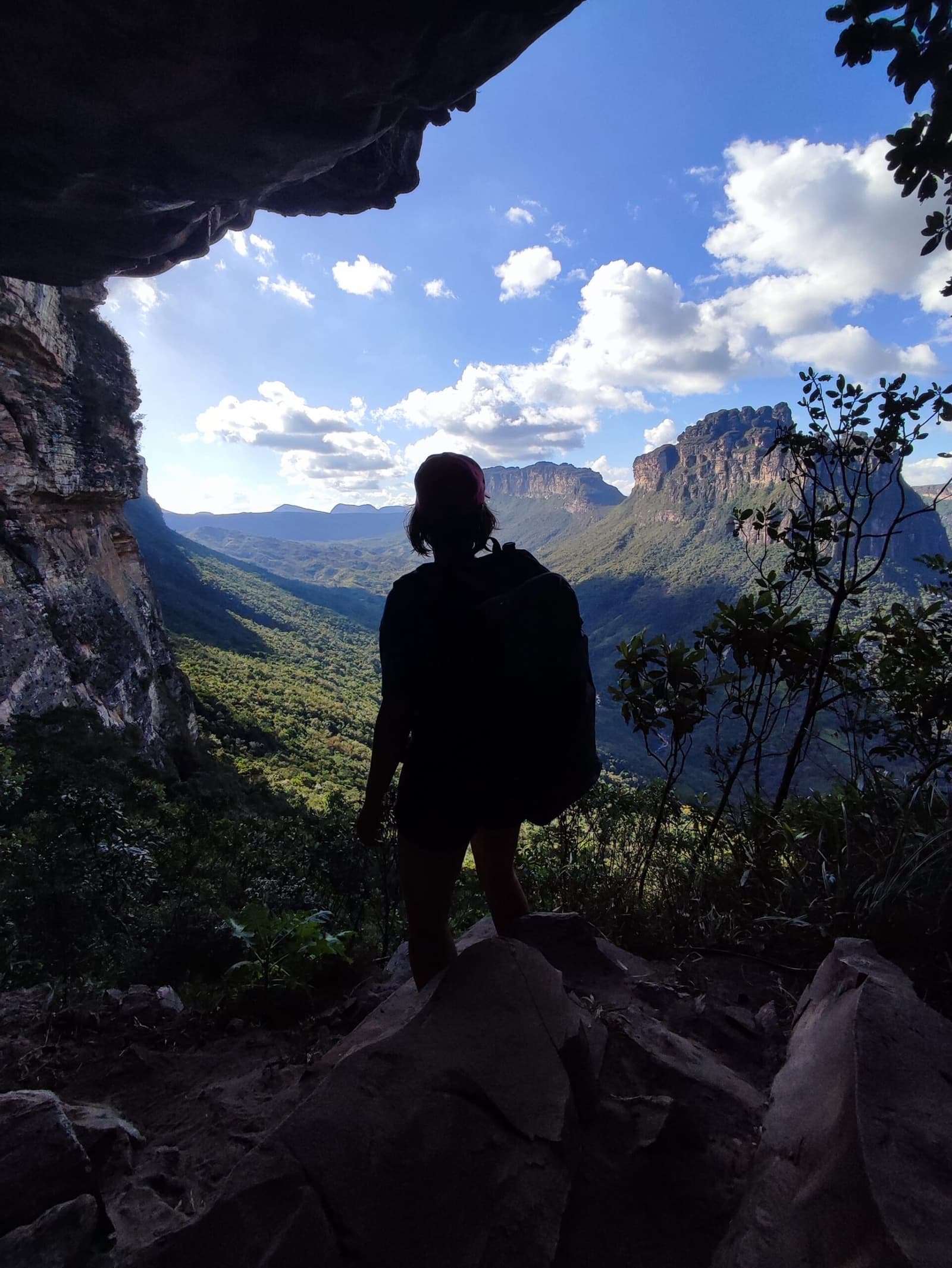 That moment when the cave frames everything and you just stop. A hiker's silhouette stands beneath a rocky overhang on the Pati Valley hike, gazing out over a sweeping green canyon and flat-topped sandstone escarpments under a blue sky