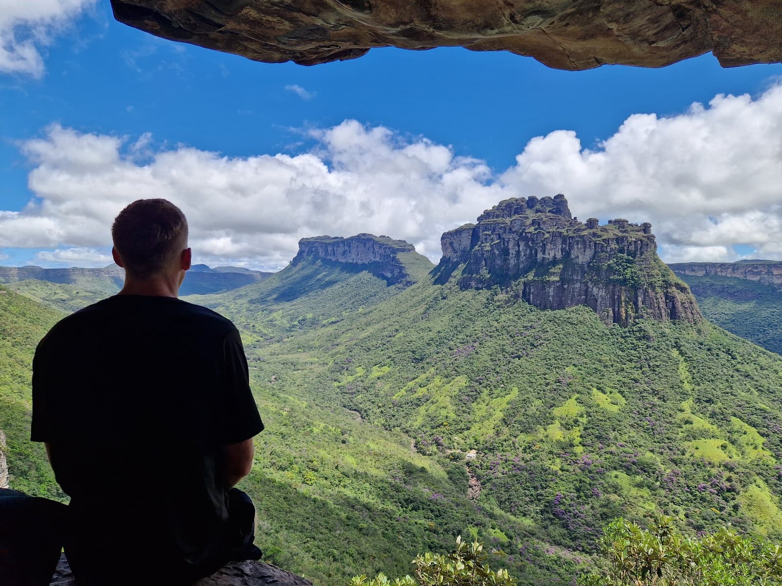 A frame nature built, a view you have to earn. A hiker sits inside a rocky cave overhang gazing out at the dramatic sandstone mesa of Morro do Castelo rising above lush green hills on the Pati Valley hike