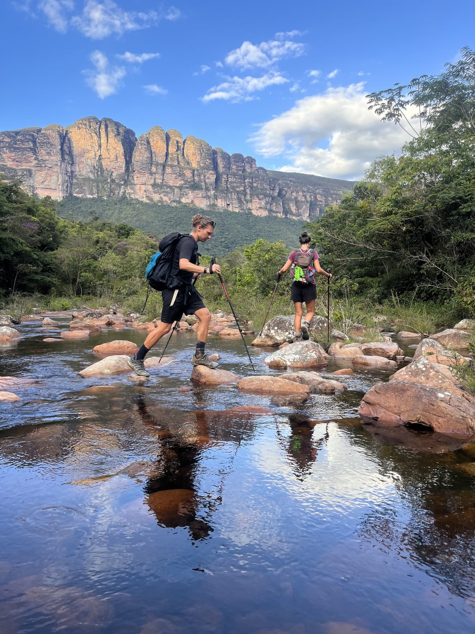 The river crossing is where trekking poles earn their keep. Two hikers with trekking poles stepping across rounded rocks in a shallow river on the Pati Valley hike, with dramatic sandstone cliffs rising behind the green forest under a blue sky