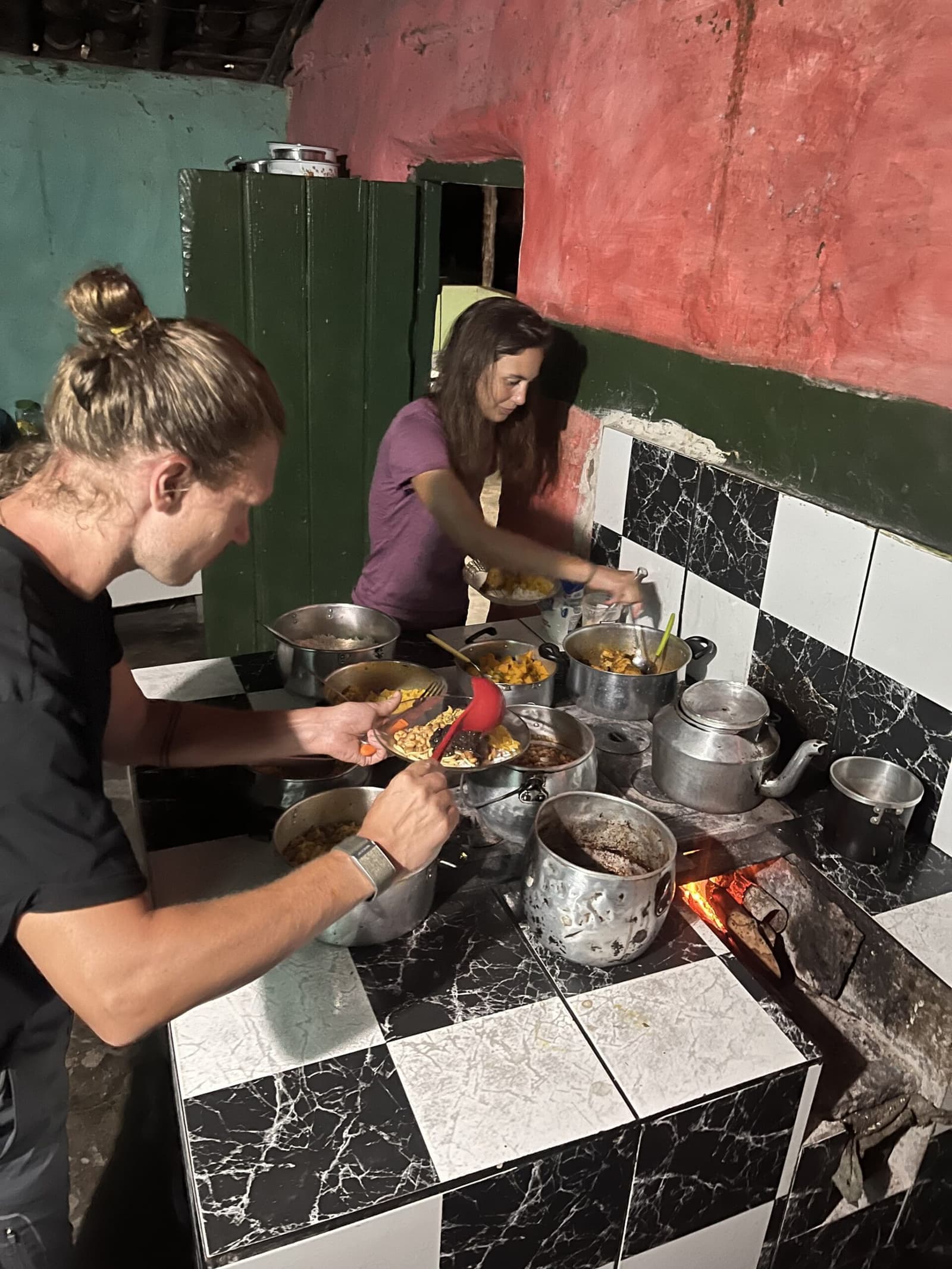 Hot food tastes better when the fire is real and the day was long. Two travelers serving themselves from multiple pots of hot food on a tiled wood-fire stove during a Pati Valley hike homestay dinner