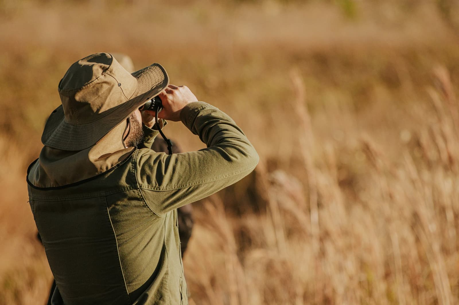 The grass is high, the list is long, and patience is its own reward. A bearded traveller in a wide-brim hat raises binoculars to scan golden Cerrado grassland during a birdwatching excursion at Pousada Trijunção