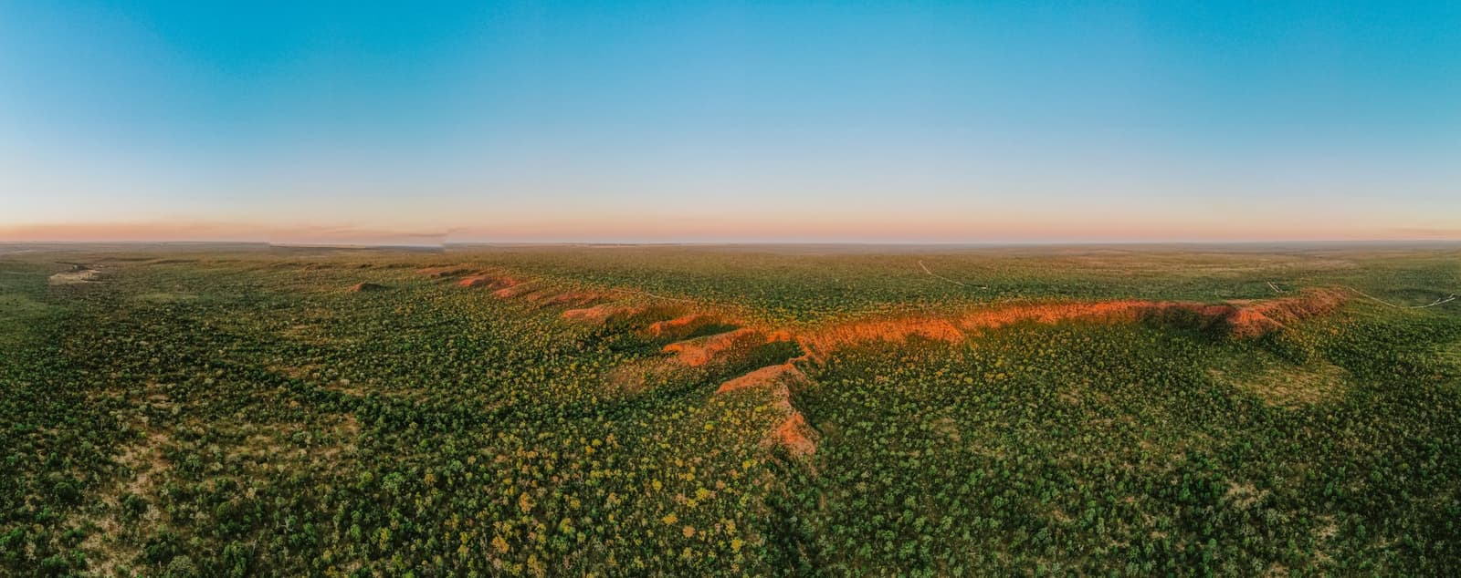 The Cerrado runs to the horizon, and that iron-red ridge reminds you the land has its own ideas about colour. Aerial panoramic view of Pousada Trijunção's vast Cerrado landscape at sunset, with a vivid red laterite ridge cutting through dense green savanna under a pastel pink and blue sky