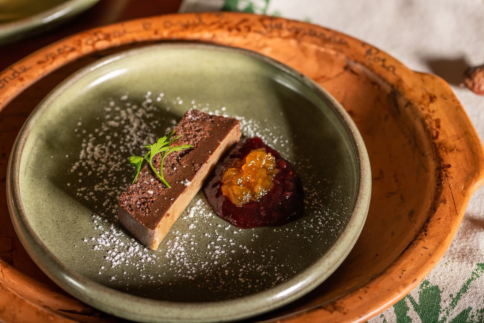 The kitchen here takes the Cerrado seriously, right down to dessert. Plated chocolate terrine slice dusted with cocoa powder and icing sugar, served alongside a jewel-toned fruit compote with golden caviar pearls on a sage-green ceramic plate at Pousada Trijunção