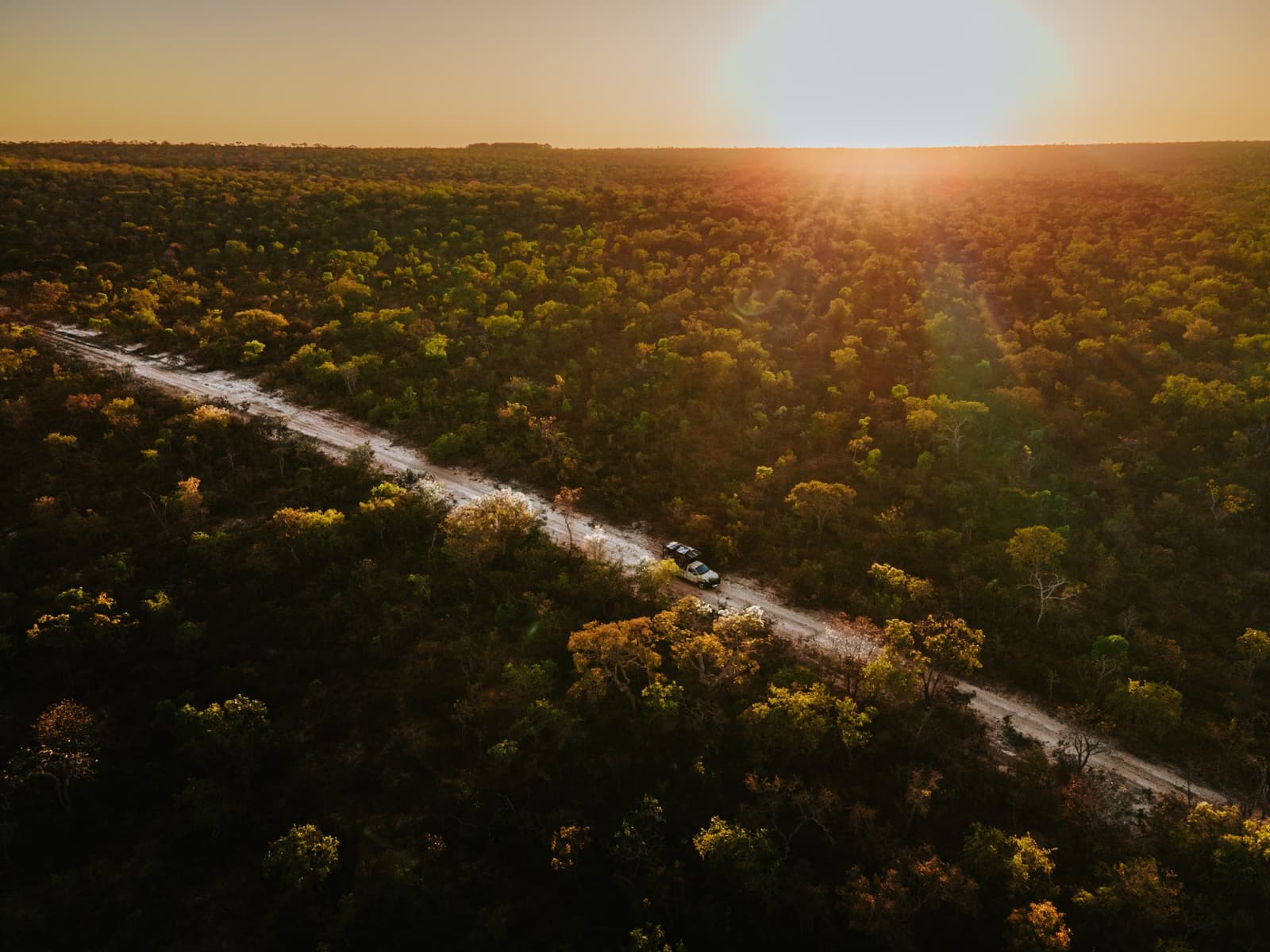The road ahead, the canopy all around, and the sun doing its best work. Aerial view of two 4x4 vehicles on a sandy dirt road cutting through dense Cerrado woodland at Pousada Trijunção, bathed in golden sunset light