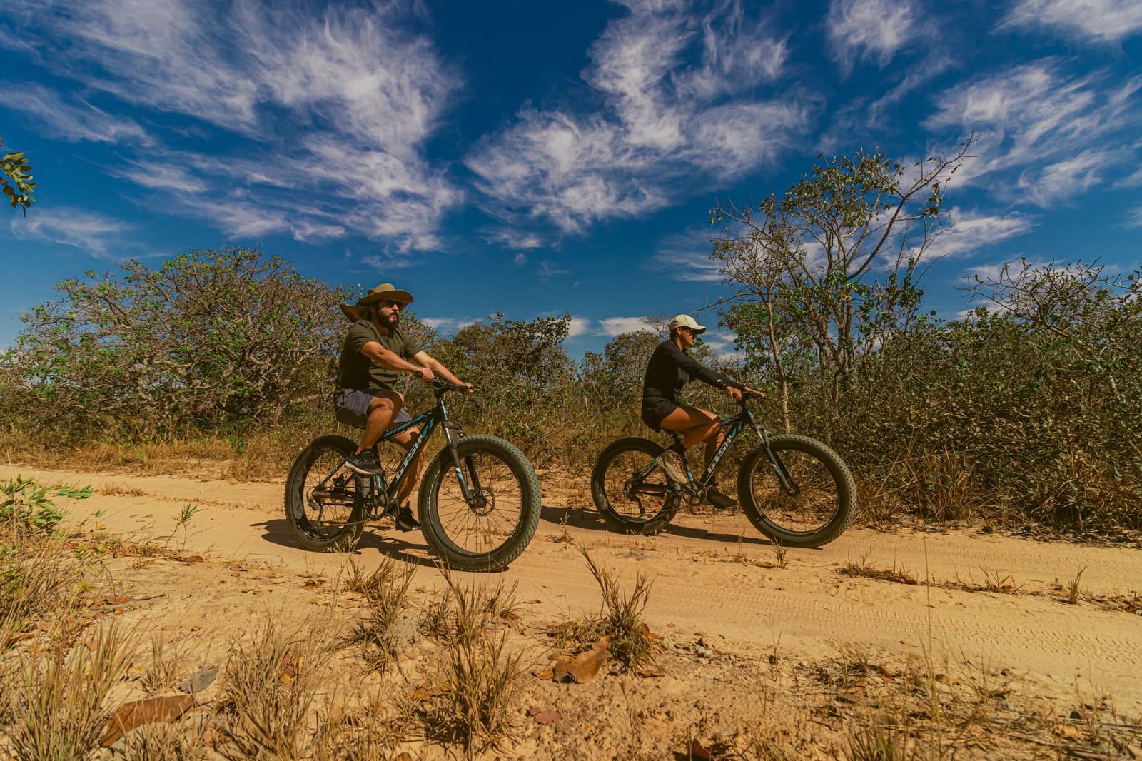 Fat tires were made for sand. The Cerrado makes you work for the view. Two cyclists riding fat bikes along a sandy Cerrado trail at Pousada Trijunção, surrounded by native cerrado shrubs under a vivid blue sky