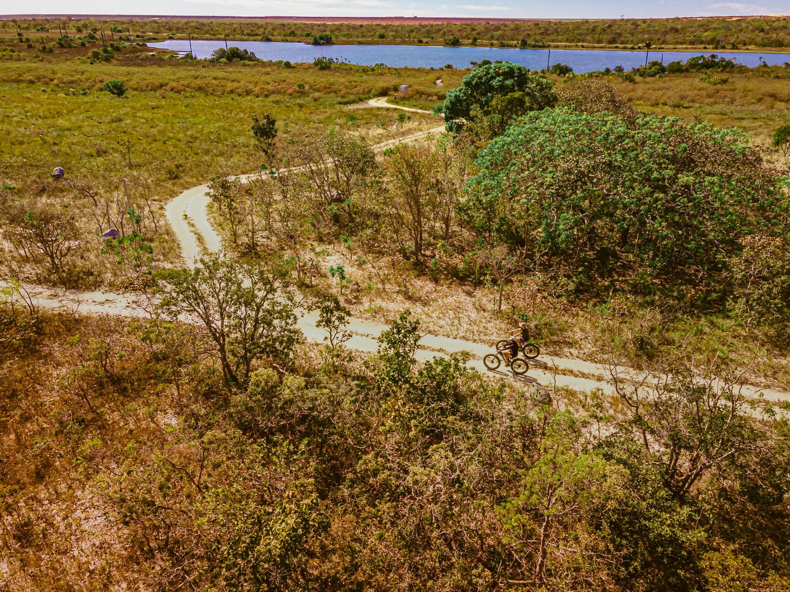 The trail picks its way through the scrub, and the lake is always just ahead. Aerial view of two cyclists riding fat bikes along a winding dirt trail through Cerrado scrubland at Pousada Trijunção, with a blue lagoon visible in the background