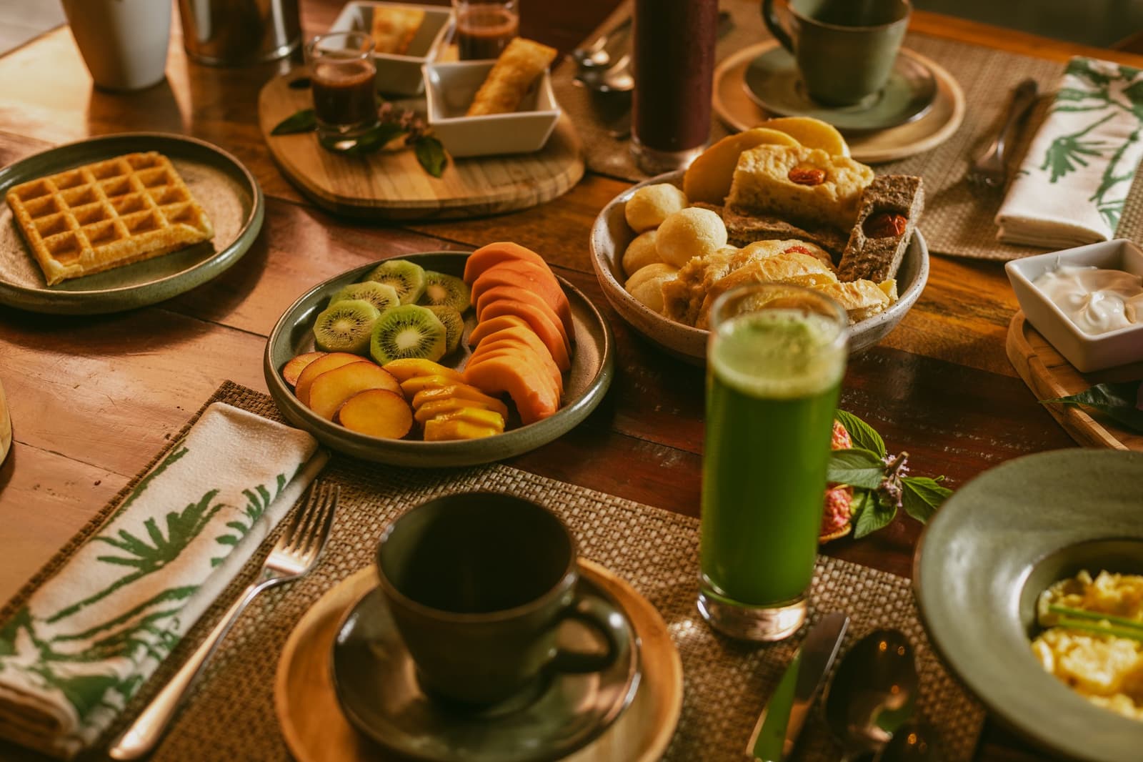 The kind of breakfast that makes you forget you have a game drive in an hour. A generous Pousada Trijunção breakfast table laid with sliced kiwi, peach and papaya, golden waffles, warm bread rolls, a vibrant green juice and a ceramic coffee mug on a woven placemat