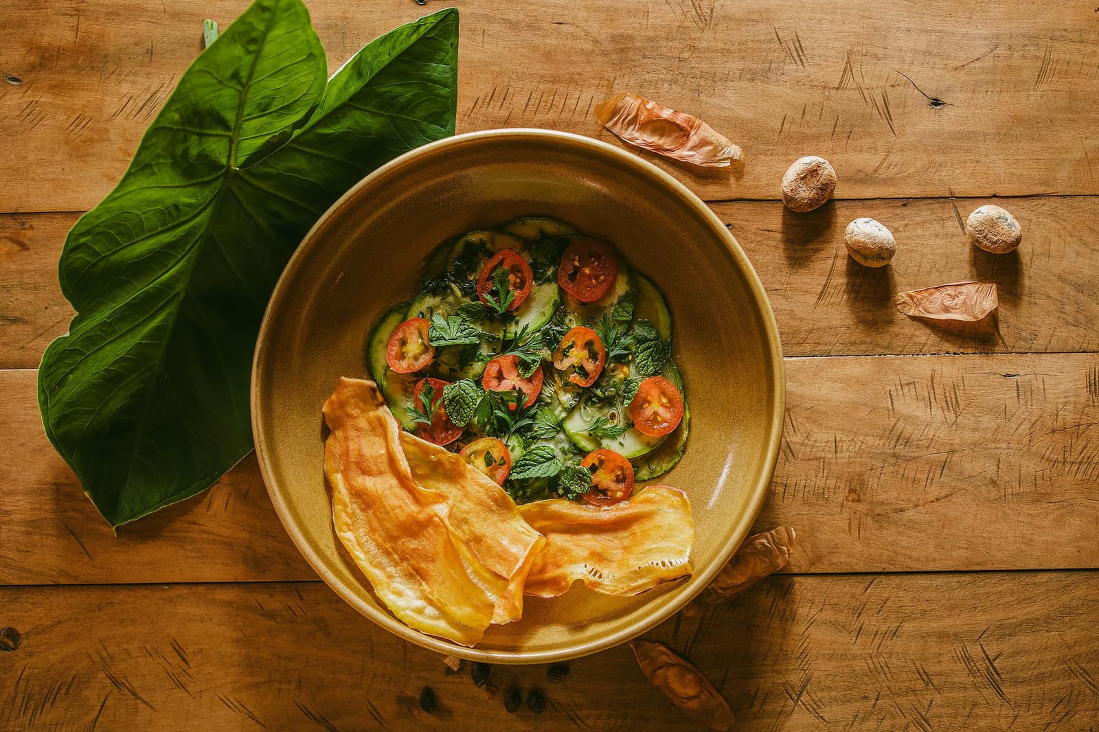 Lodge cooking that tastes exactly like where you are. Overhead view of a ceramic bowl filled with sliced zucchini, cherry tomatoes and fresh herbs served with crispy chips at Pousada Trijunção, surrounded by tropical leaves and wild seeds on a rustic wooden table