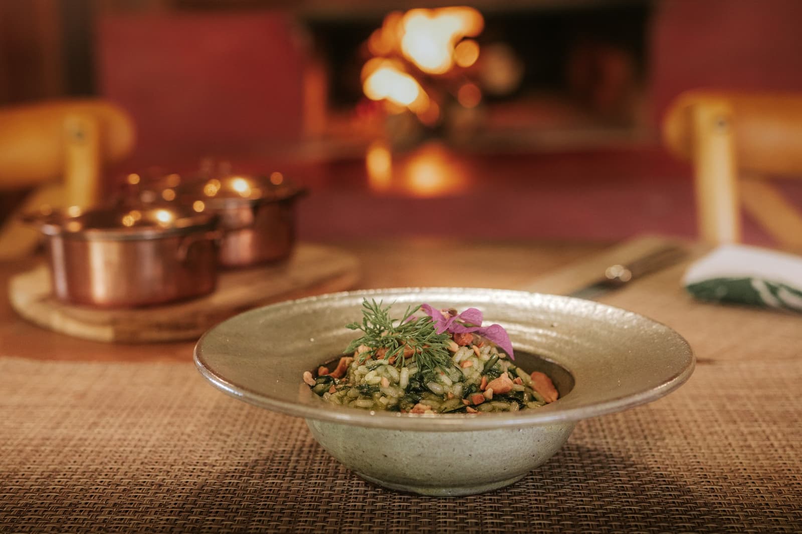 Dinner tastes better when the fireplace is doing the heavy lifting. A handcrafted ceramic bowl of herb risotto garnished with fresh dill and a pink edible flower, served fireside at Pousada Trijunção with copper pots glowing in the warm background
