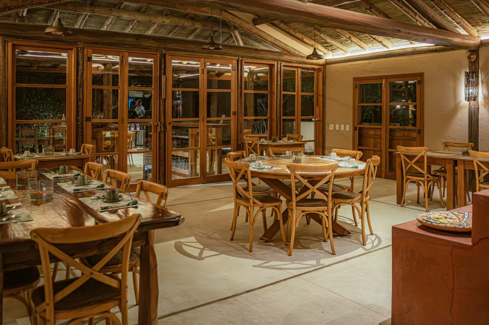 Good food tastes even better when the ceiling is made of straw. Warmly lit dining room at Pousada Trijunção featuring rustic wooden cross-back chairs, round and long communal tables set with green napkins, thatched ceiling with exposed timber beams, and large wood-framed glass doors opening to the garden